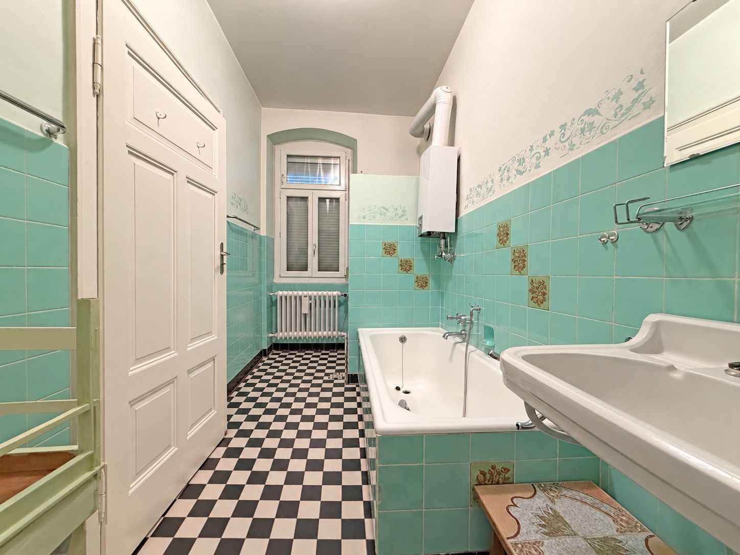 Helles Bad mit Badewanne, Waschbecken und grün gefliesten Wänden, kariertem Boden sowie Fenster am Ende.