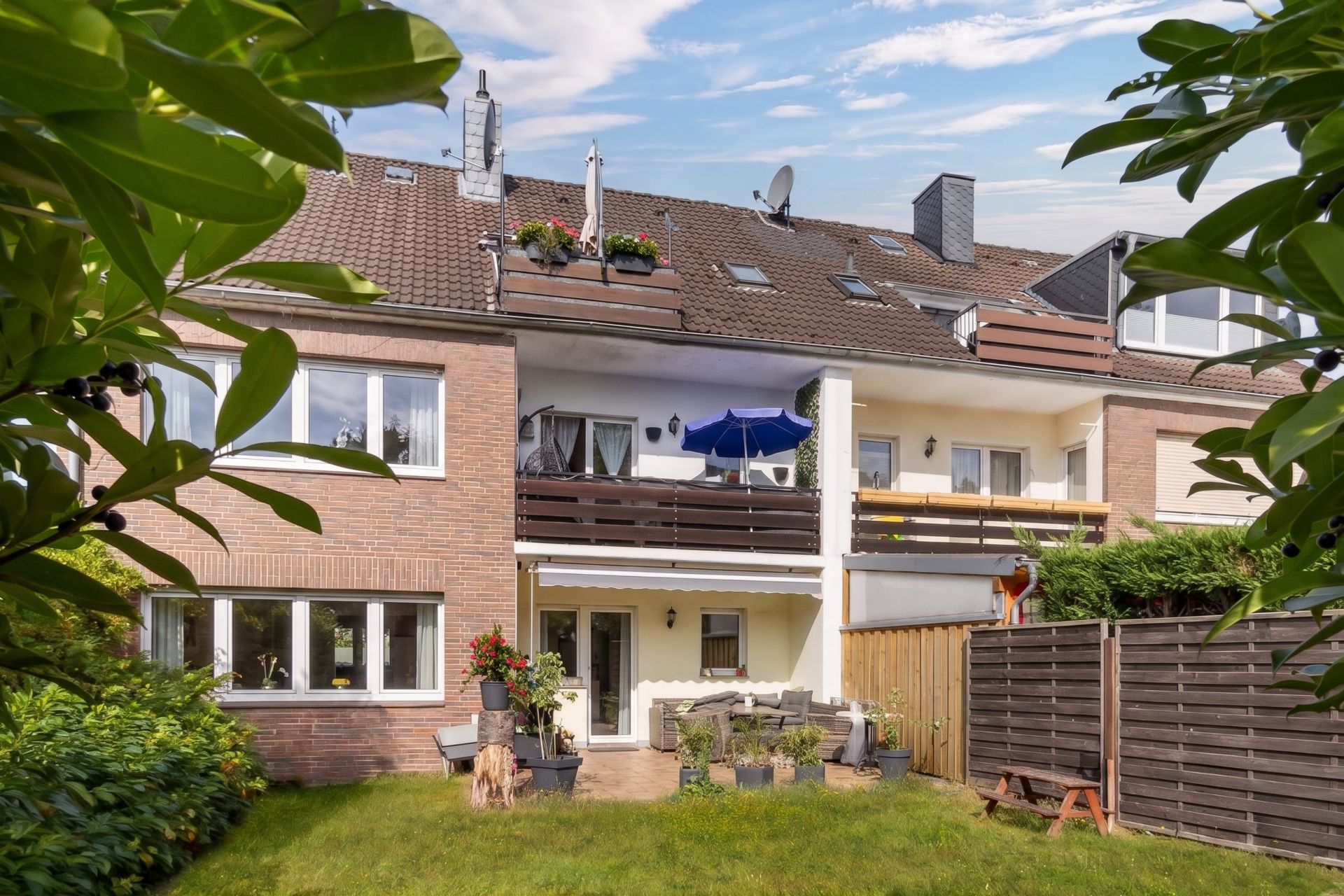 Rückseite eines Reihenhauses mit Balkon, Terrasse und kleinem Garten, umgeben von Pflanzen.