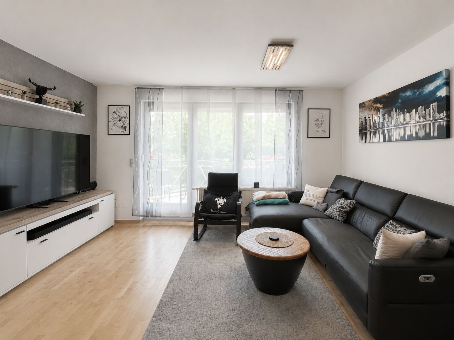 Modernes Wohnzimmer mit schwarzem Sofa, Couchtisch, TV und großen Fenstern mit transparenten Vorhängen.