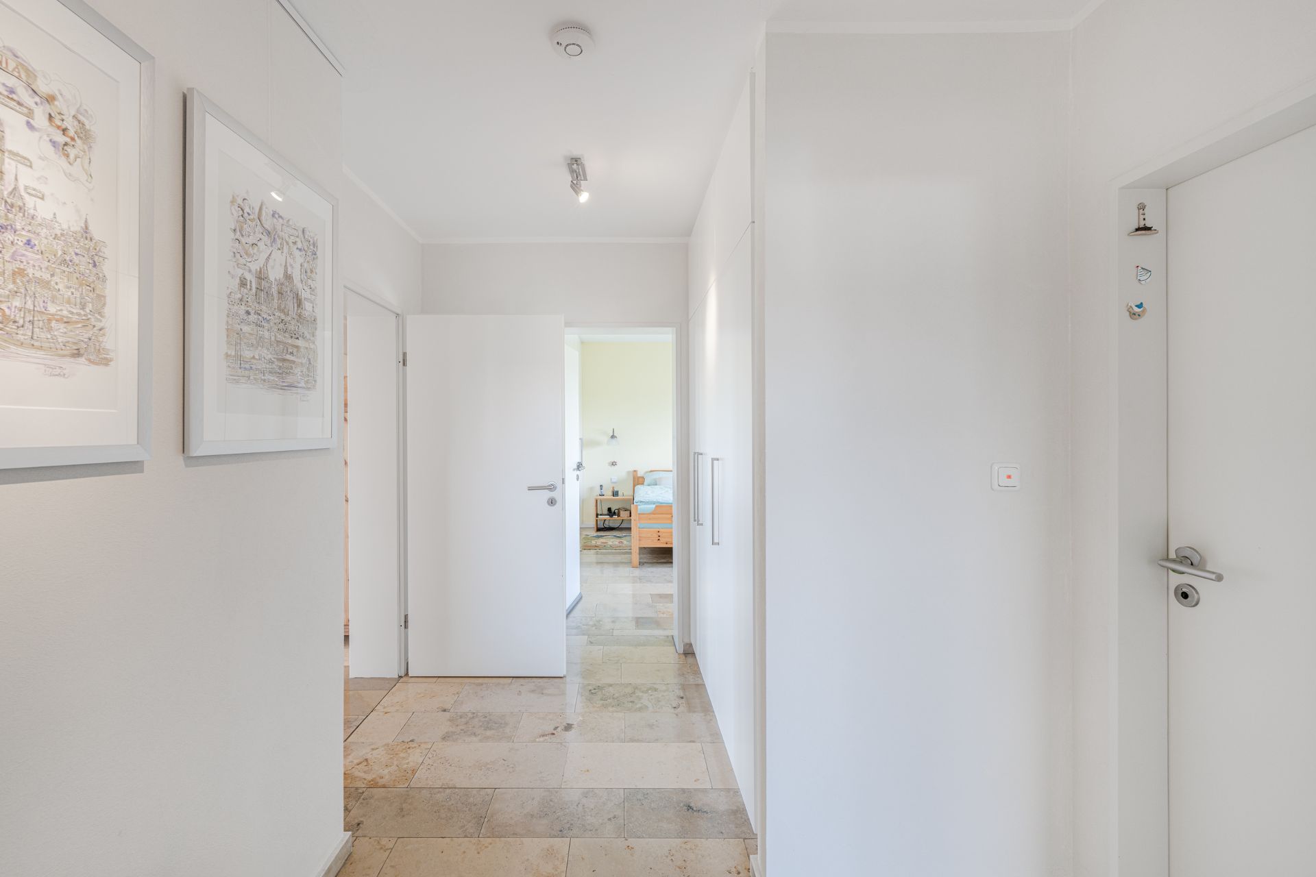 Heller Flur mit weißen Türen, Bildern an der Wand und Blick in ein Zimmer mit Holzbett.