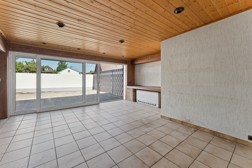Heller Raum mit großen Glas-Schiebetüren, Fliesenboden und Holzdecke, Blick auf Terrasse.