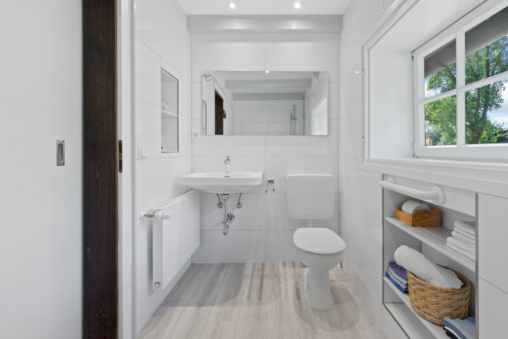 Helles Bad mit WC, Waschbecken, Spiegel, Fenster und Regal mit Handtüchern.