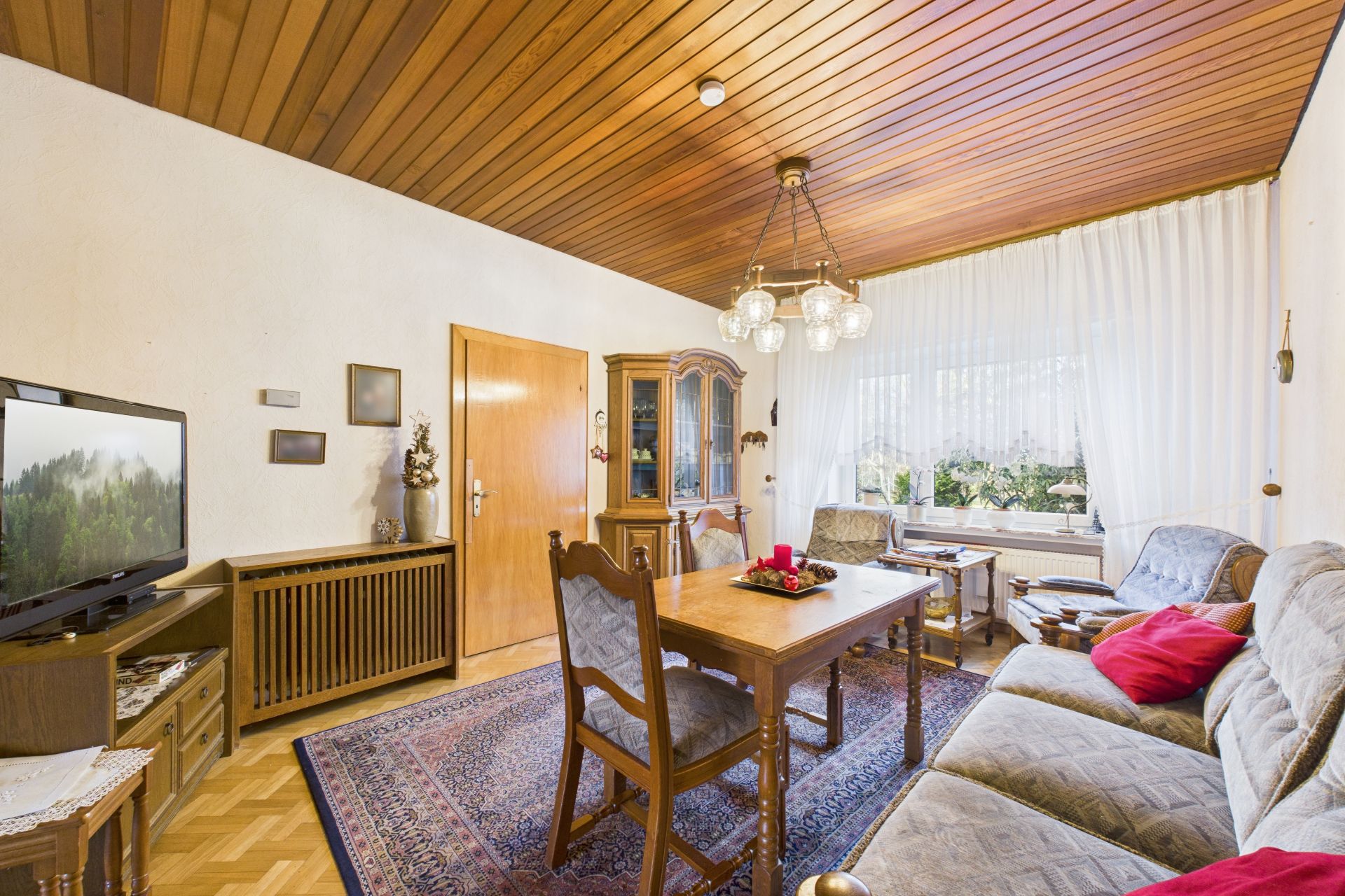 Wohnzimmer mit Holzboden, Holzbalkendecke, Sofa, Esstisch, Fernseher und großem Fenster mit Gardinen.