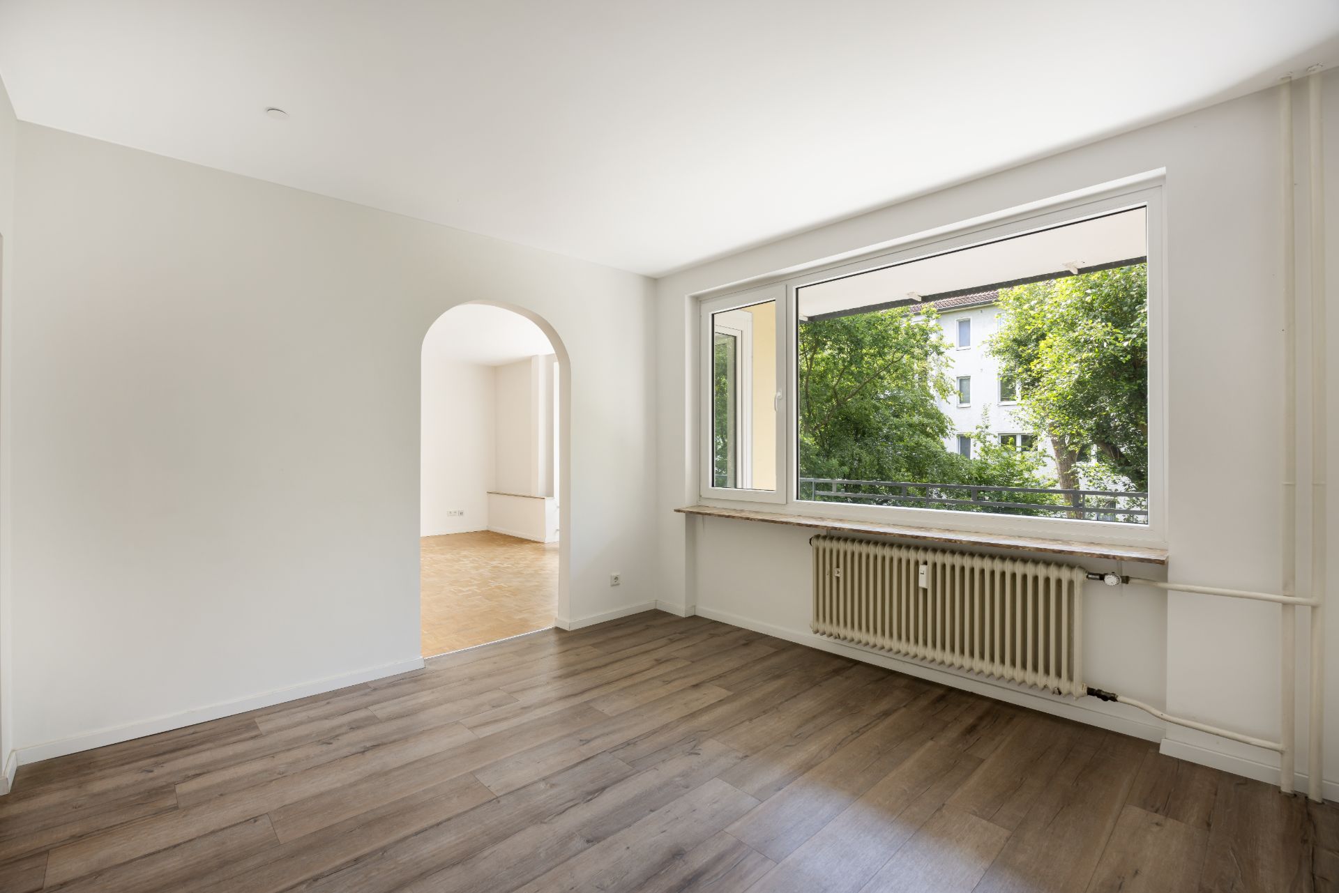 Helles Wohnzimmer mit großem Fenster, Holzboden und Blick auf grüne Bäume, Durchgang zu weiterem Raum.