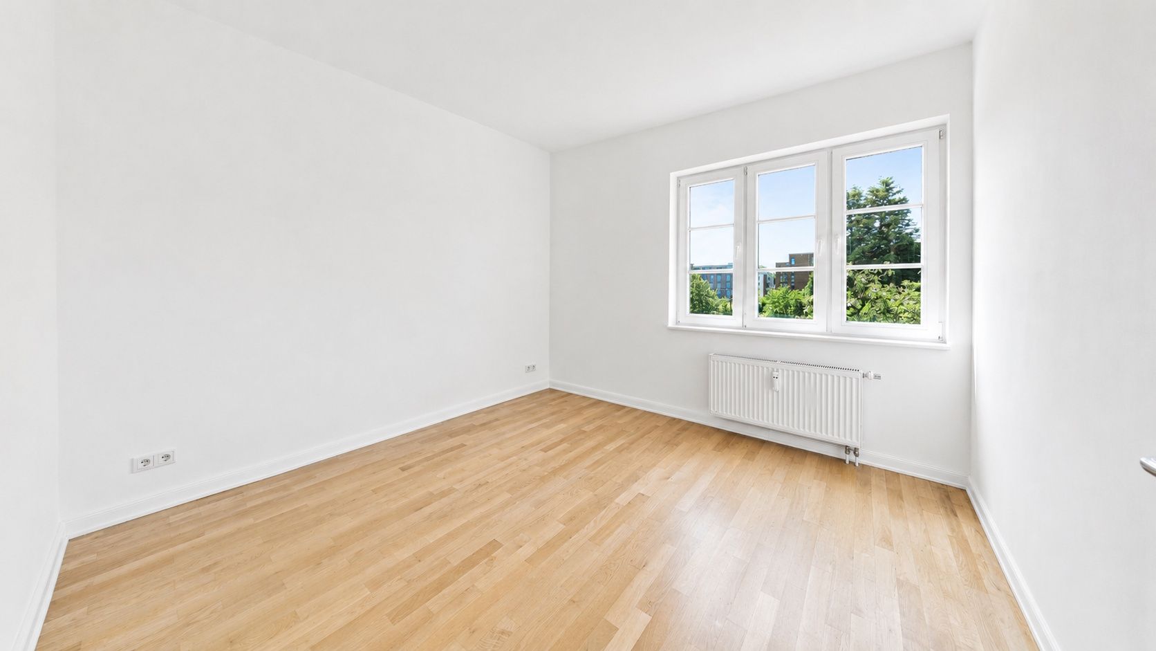 Helles, leeres Zimmer mit Parkettboden und großem Fenster mit Blick auf grüne Bäume.