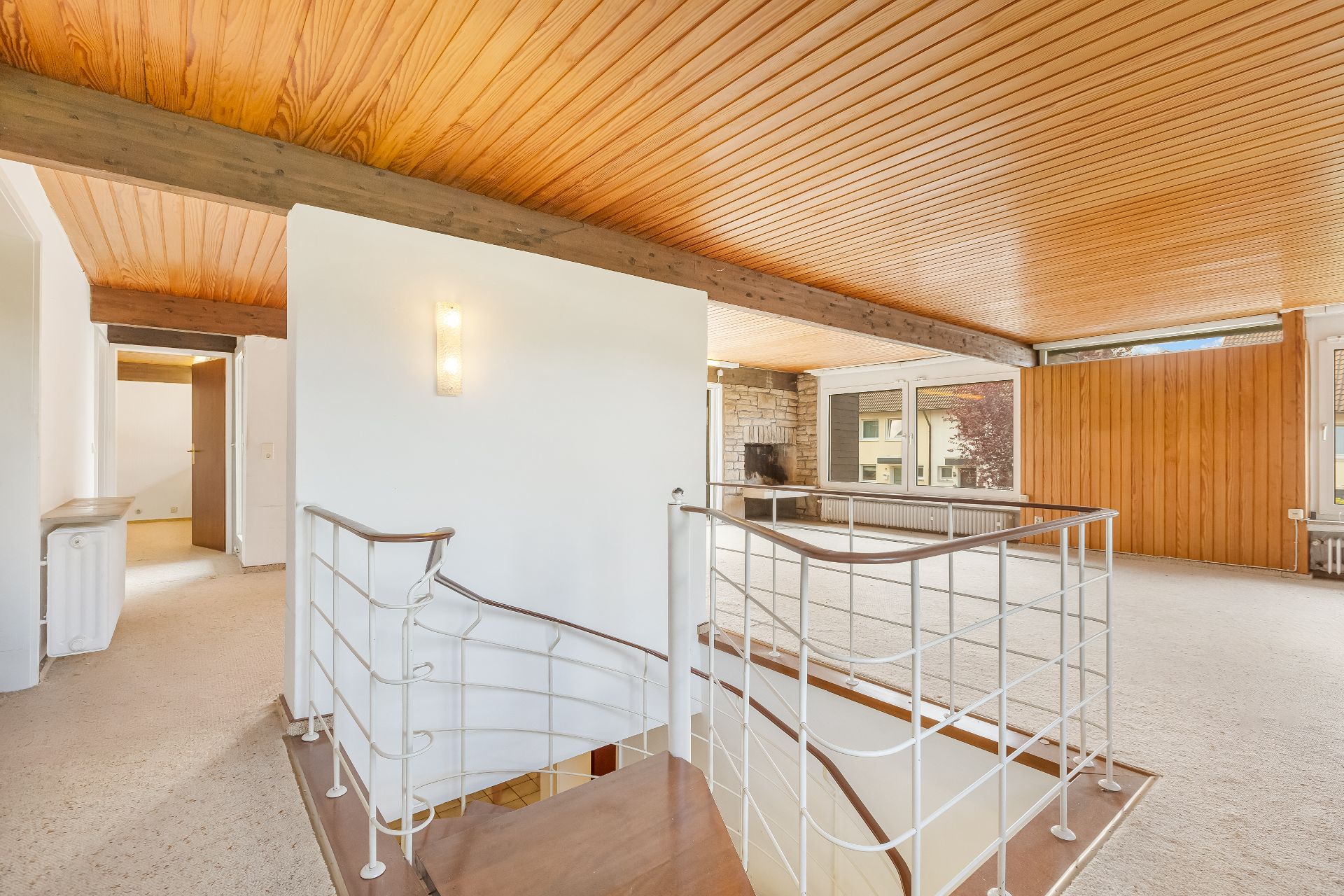 Helles Wohnzimmer mit Holzdecke, Kamin, großen Fenstern und Treppe nach unten.