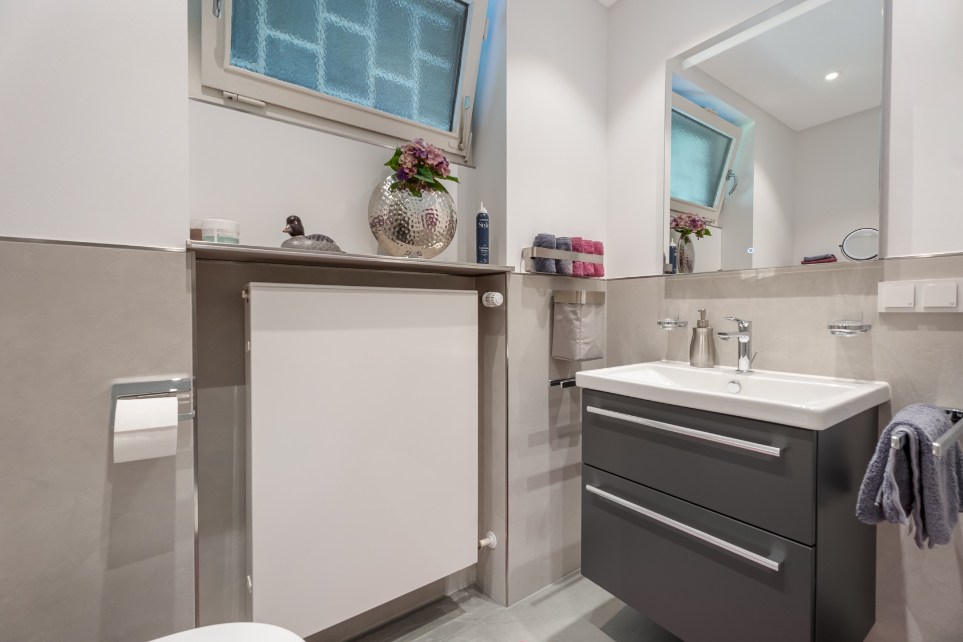 Modernes Badezimmer mit Waschbecken, Spiegel und Fenster. Rechts ist ein Handtuch zu sehen.