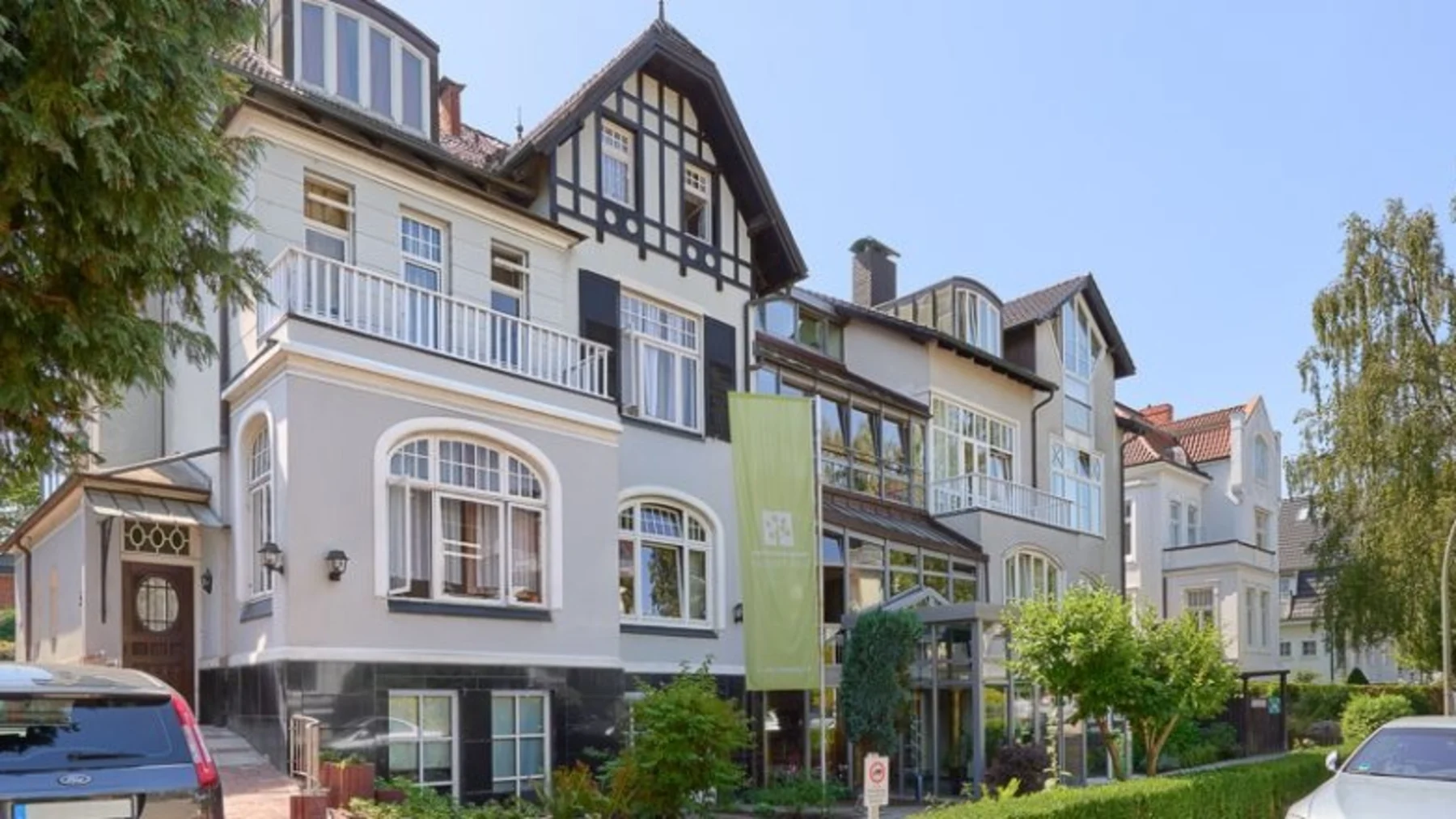 Mehrstöckiges Wohnhaus mit Erkern, Balkon und großen Fenstern, umgeben von Bäumen.