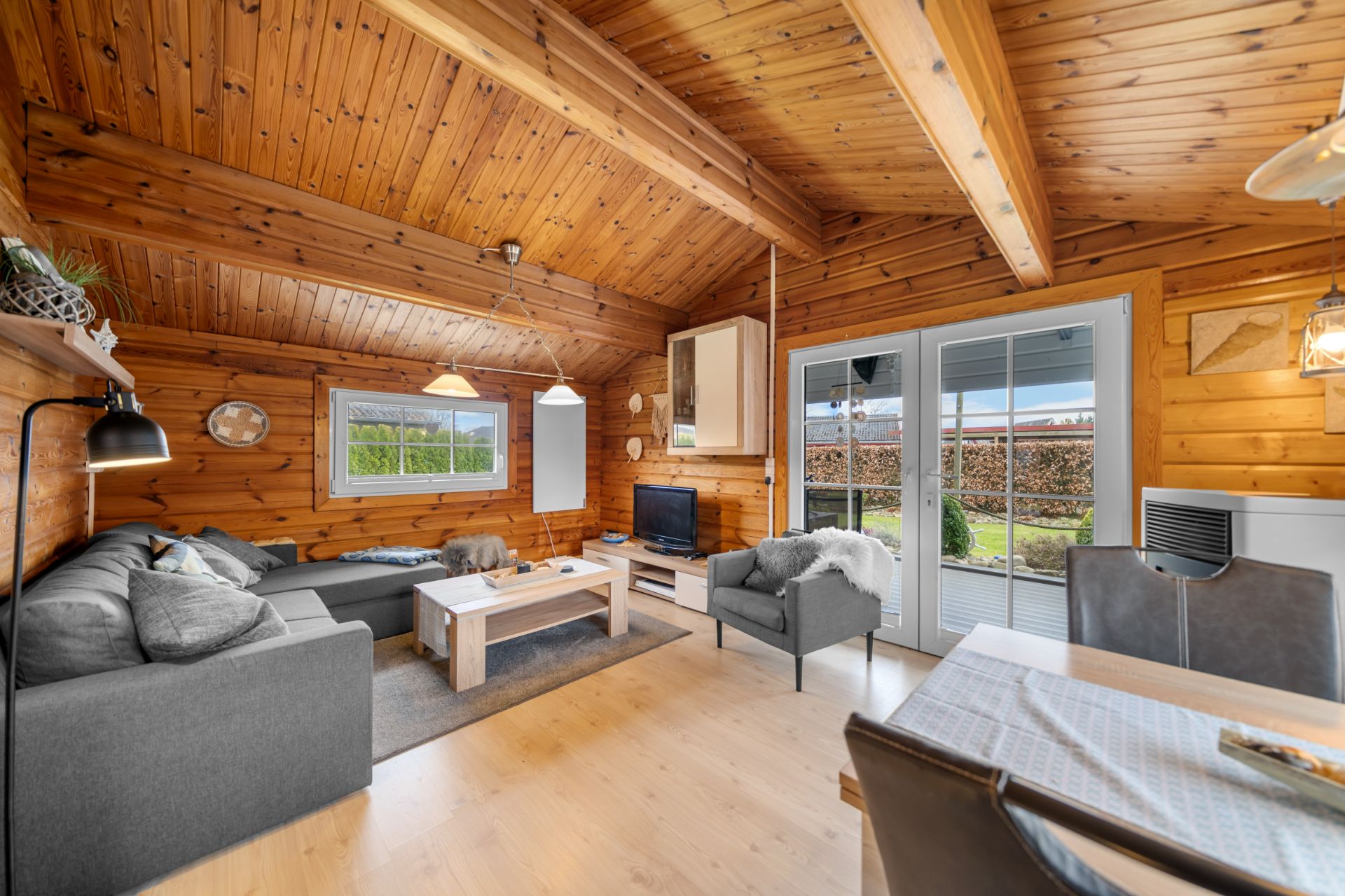 Wohnzimmer mit Holzwänden, grauem Sofa, Couchtisch, Sessel und großem Fenster mit Terrassentür.