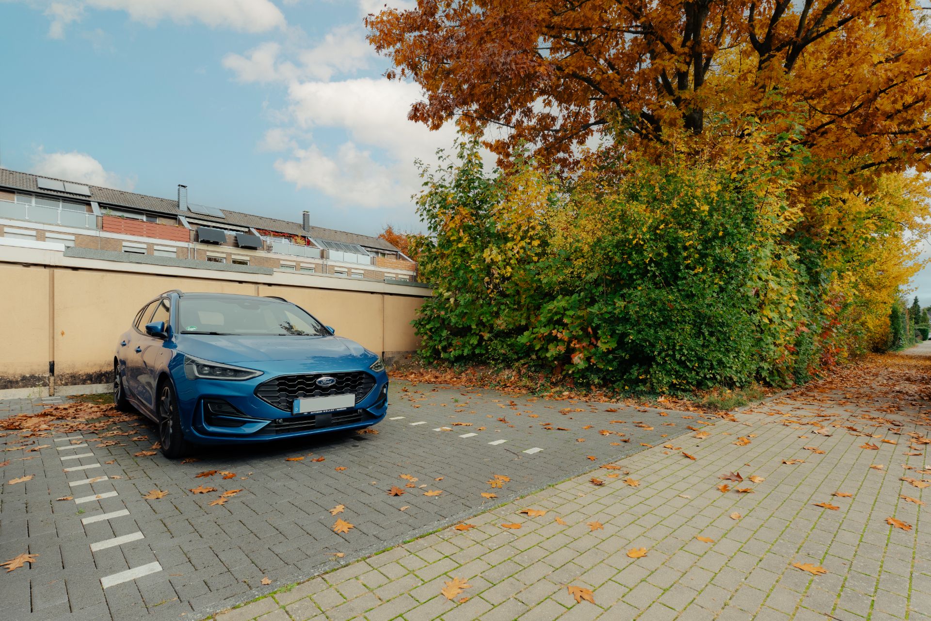 Blauer Pkw auf einem Parkplatz neben einer Hecke und herbstlichen Bäumen.