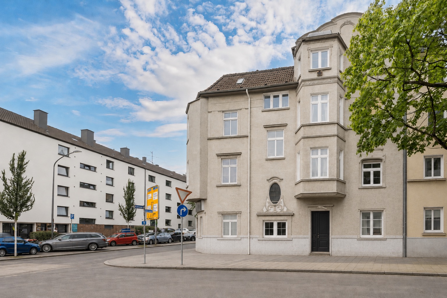 Wohnhaus an einer Straßenecke mit mehreren Fenstern und einem Baum rechts im Bild.