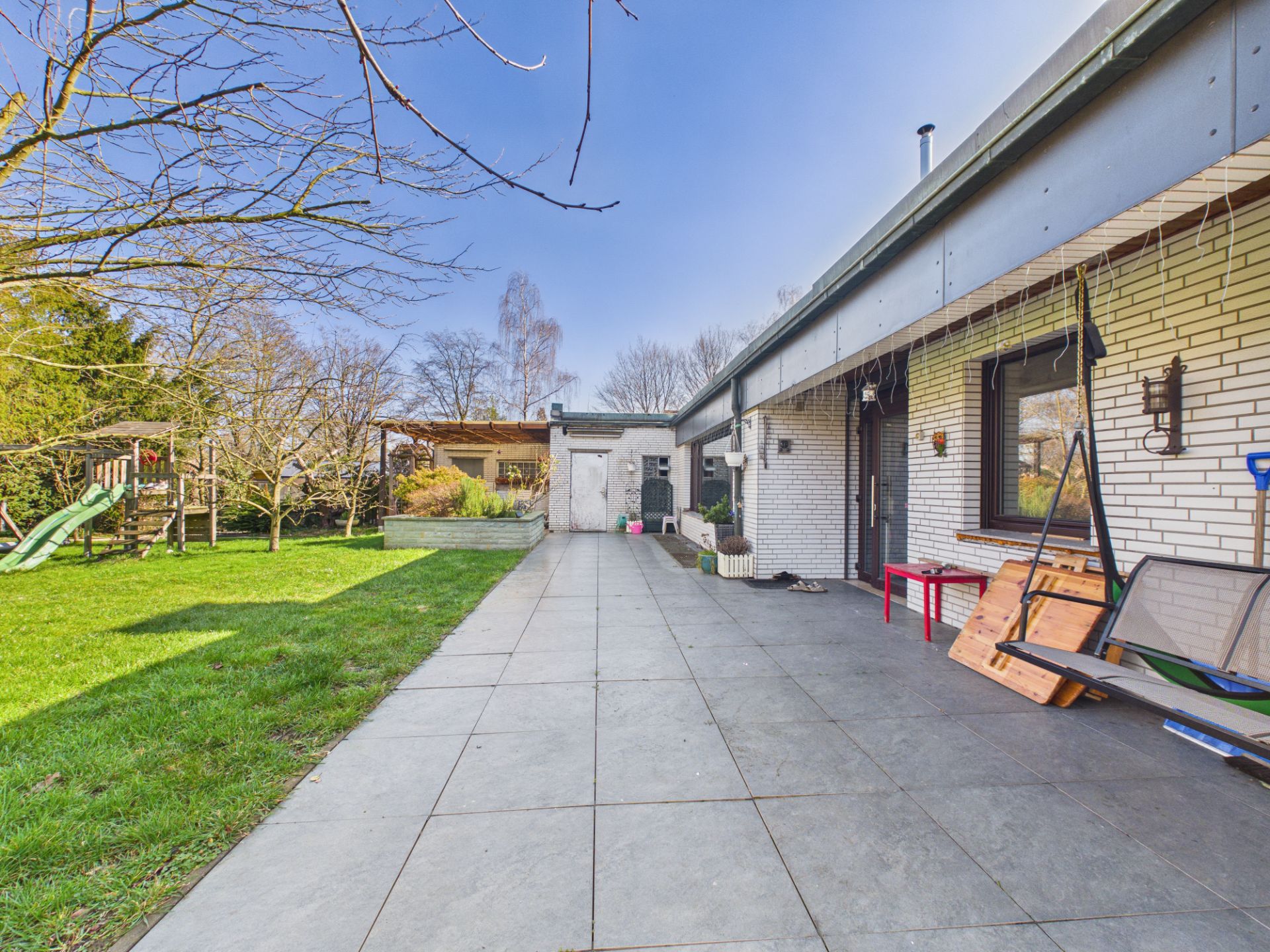 Große Terrasse neben einem weißen Haus mit Garten und Spielplatz im Hintergrund.