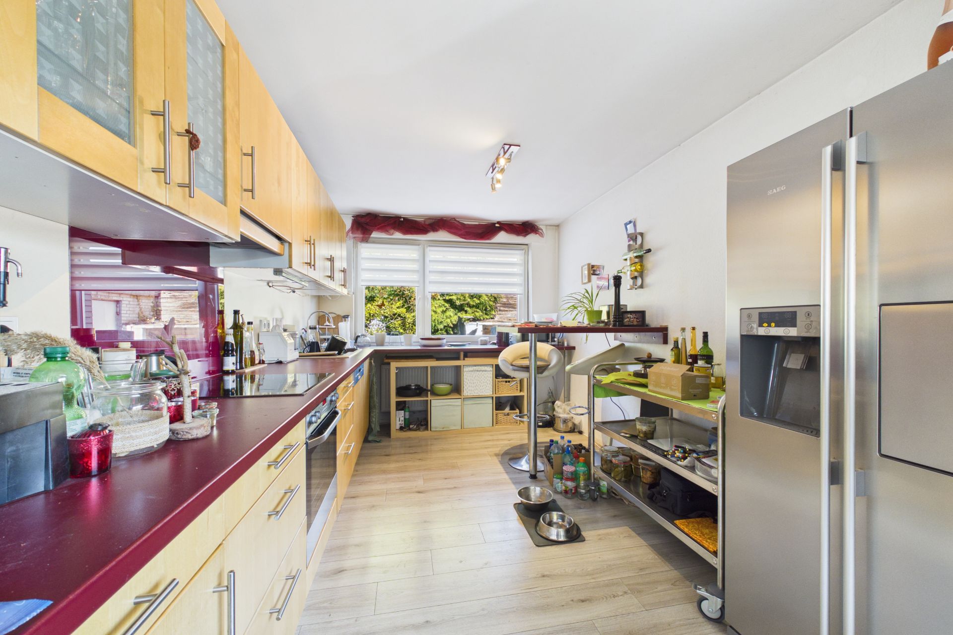Moderne Küche mit Holzmöbeln, großem Kühlschrank und Fenster; diverse Küchengeräte sichtbar.