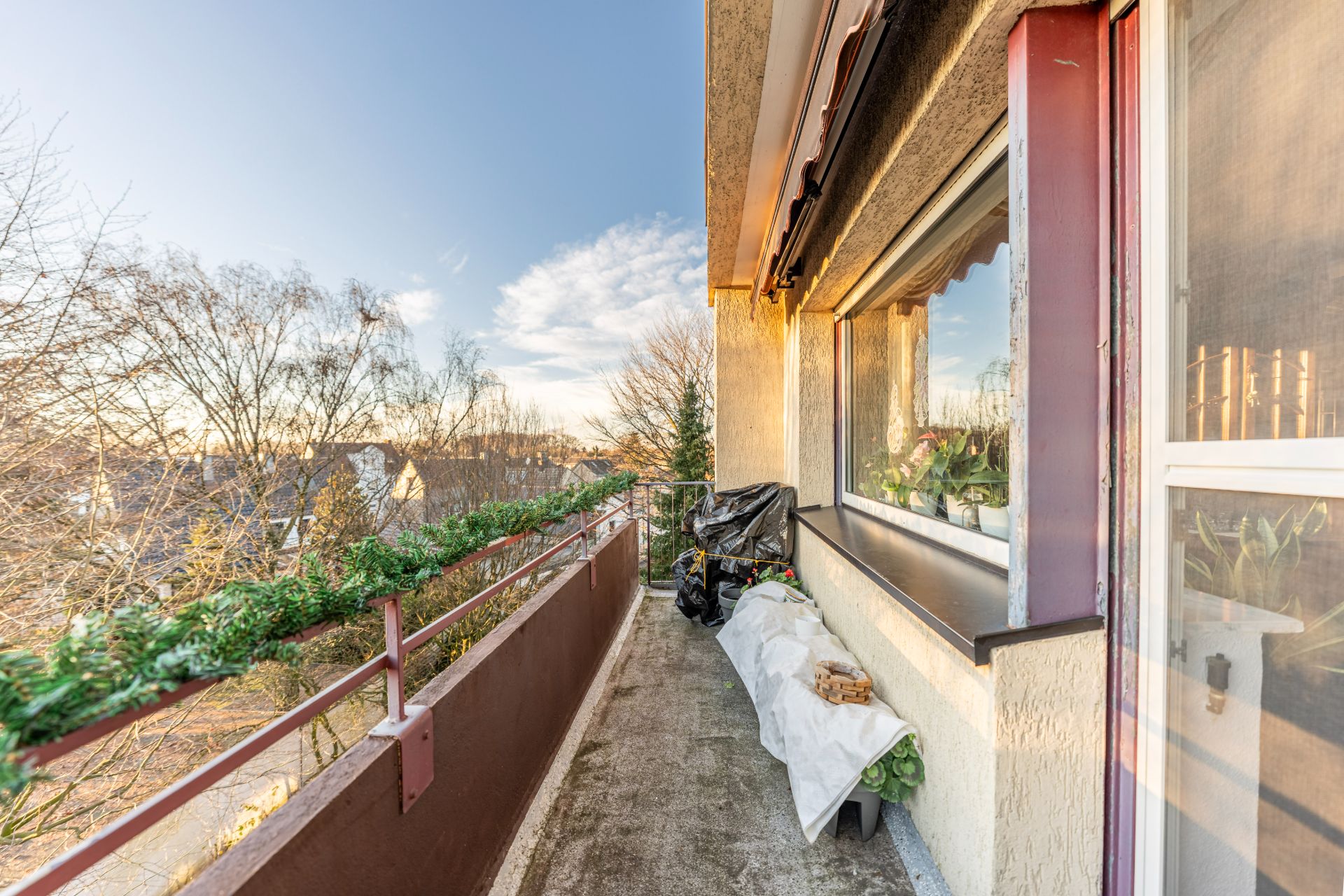 Langer Balkon mit Pflanzen auf der Brüstung und Fenster zur Wohnung, Blick auf Bäume und Häuser.