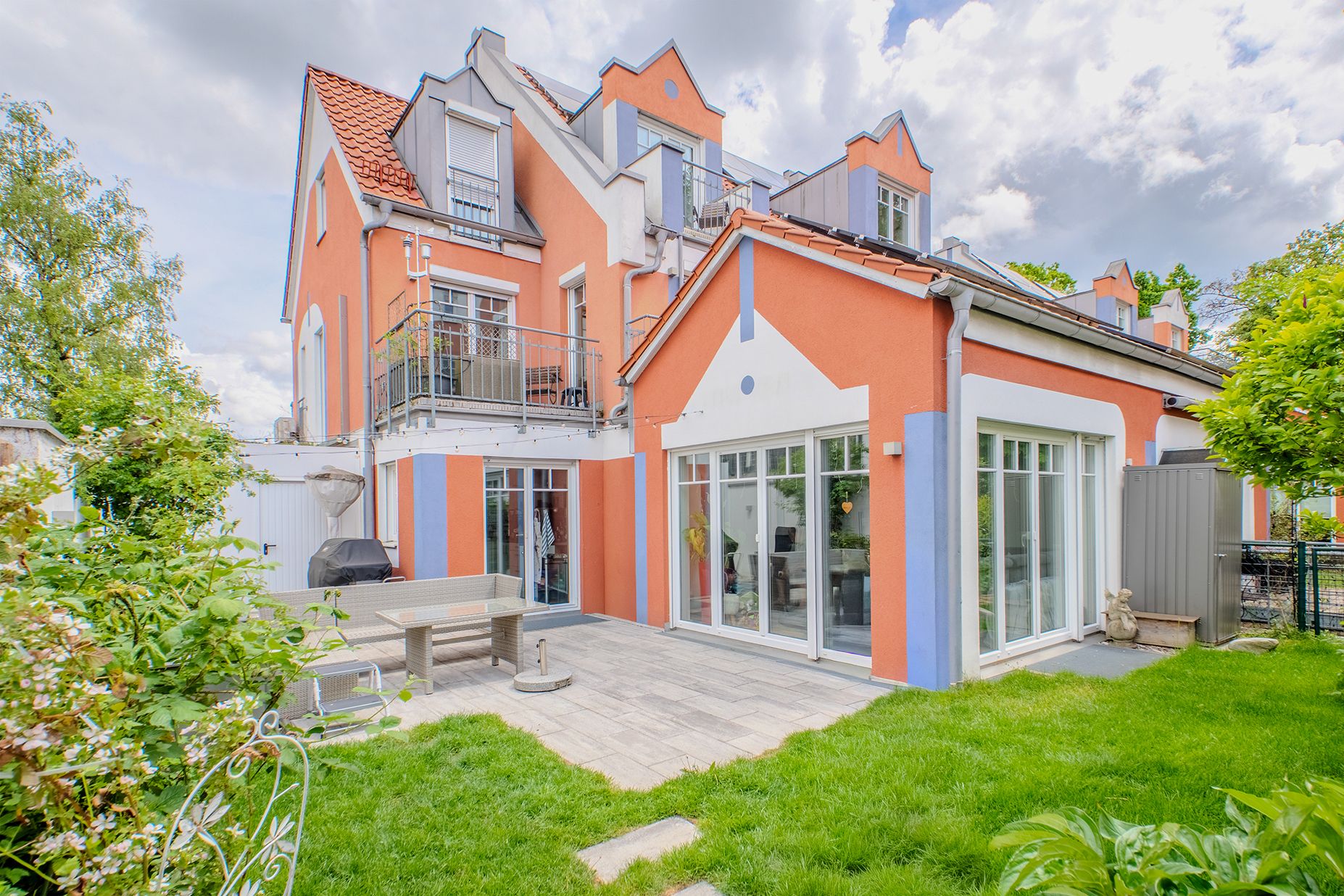 Modernes Wohnhaus mit oranger Fassade, großen Fenstern und Garten mit Terrasse und Sitzbank.
