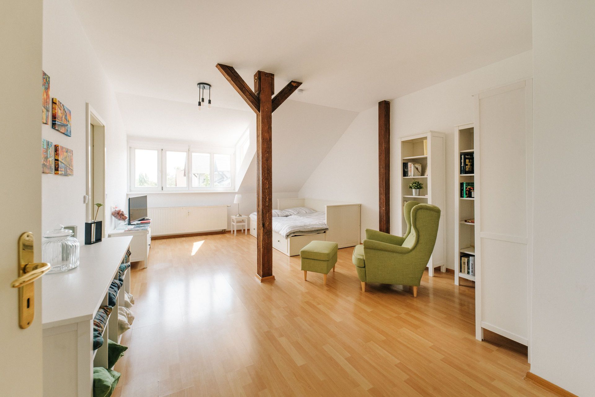 Helles Zimmer mit Holzfußboden, Bett, grünem Sessel, Bücherregalen und sichtbaren Holzbalken.
