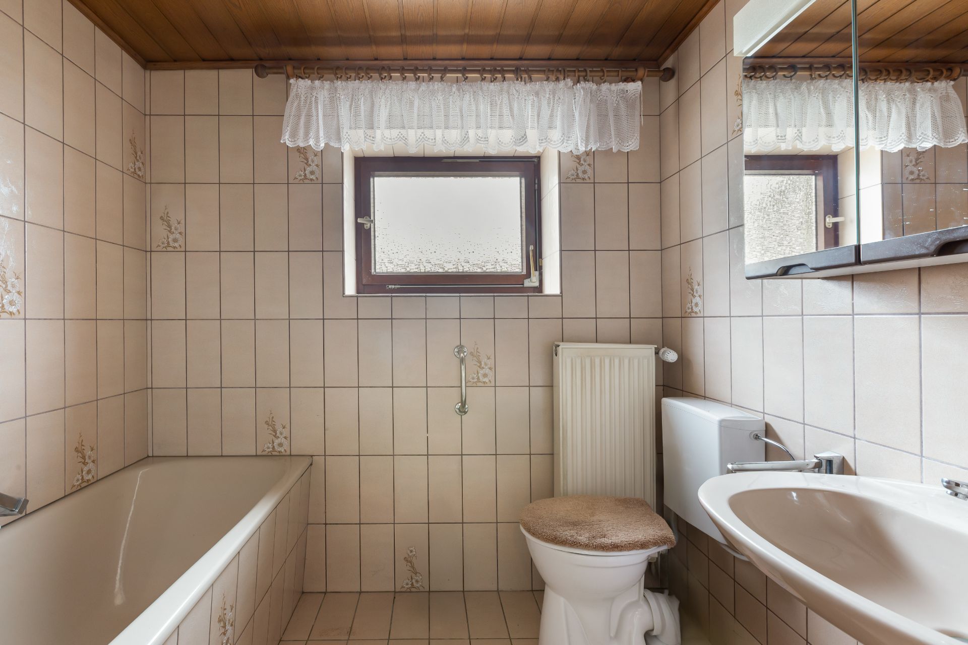 Kleines Badezimmer mit Badewanne, WC, Waschbecken und Fenster. Beige Fliesen und Holzdecke.