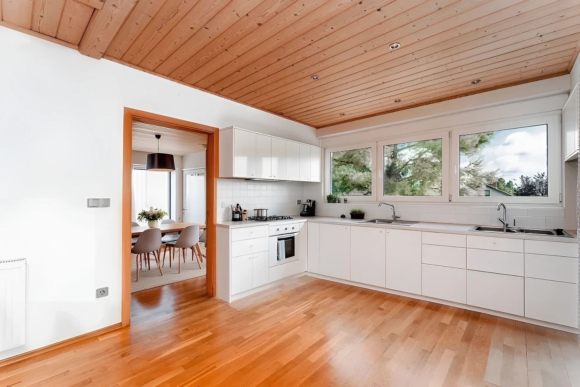 Moderne weiße Einbauküche mit Holzboden und Blick ins Esszimmer, große Fenster an der Wand.