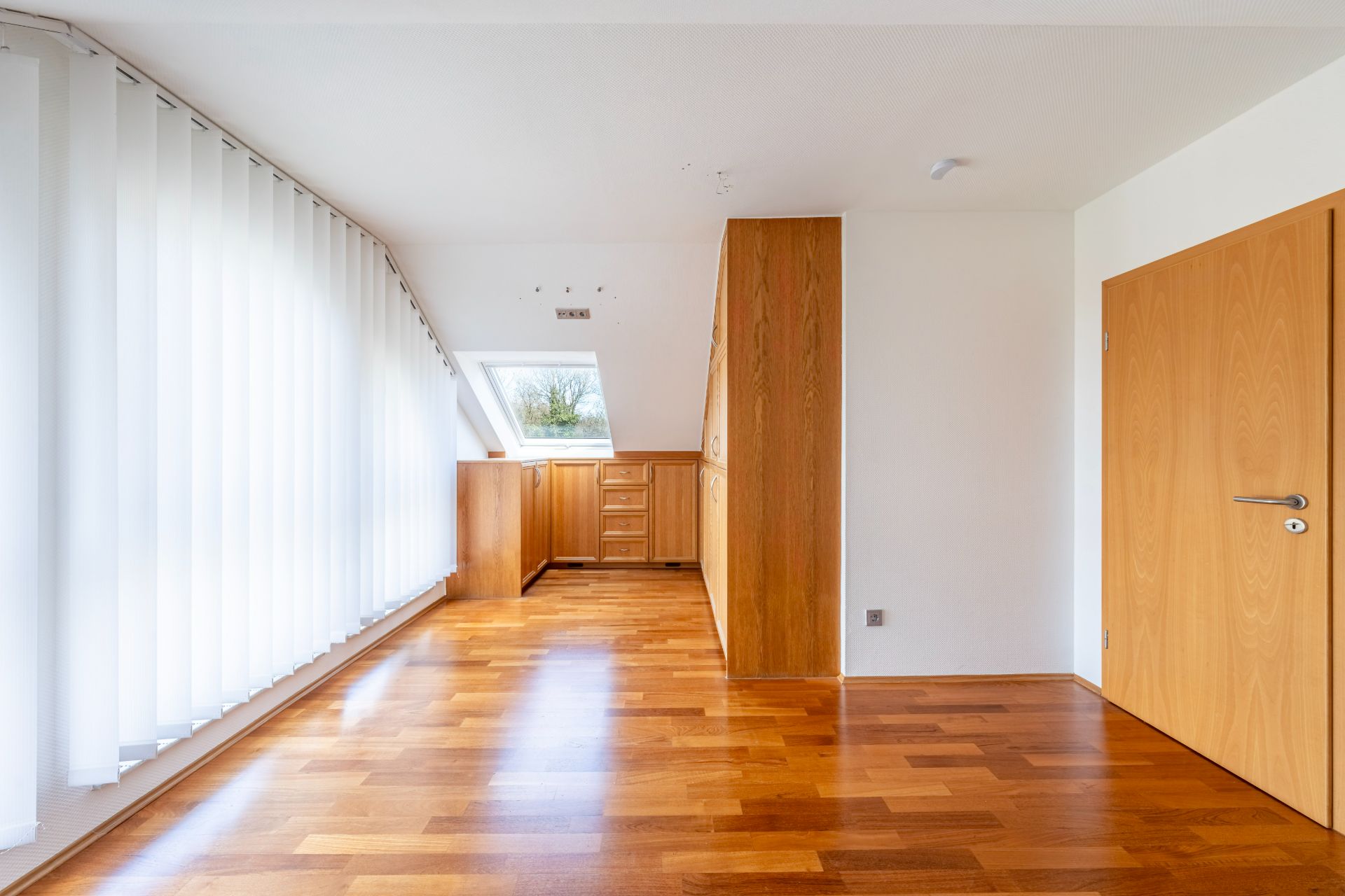 Helles Zimmer mit Parkettboden, Holzschränken und großen Fenstern mit vertikalen Jalousien.