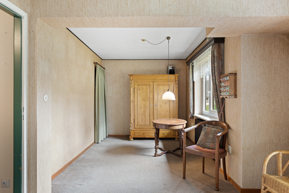 Raum mit Holzschrank, rundem Tisch, Stuhl und Fenster mit Gardine.