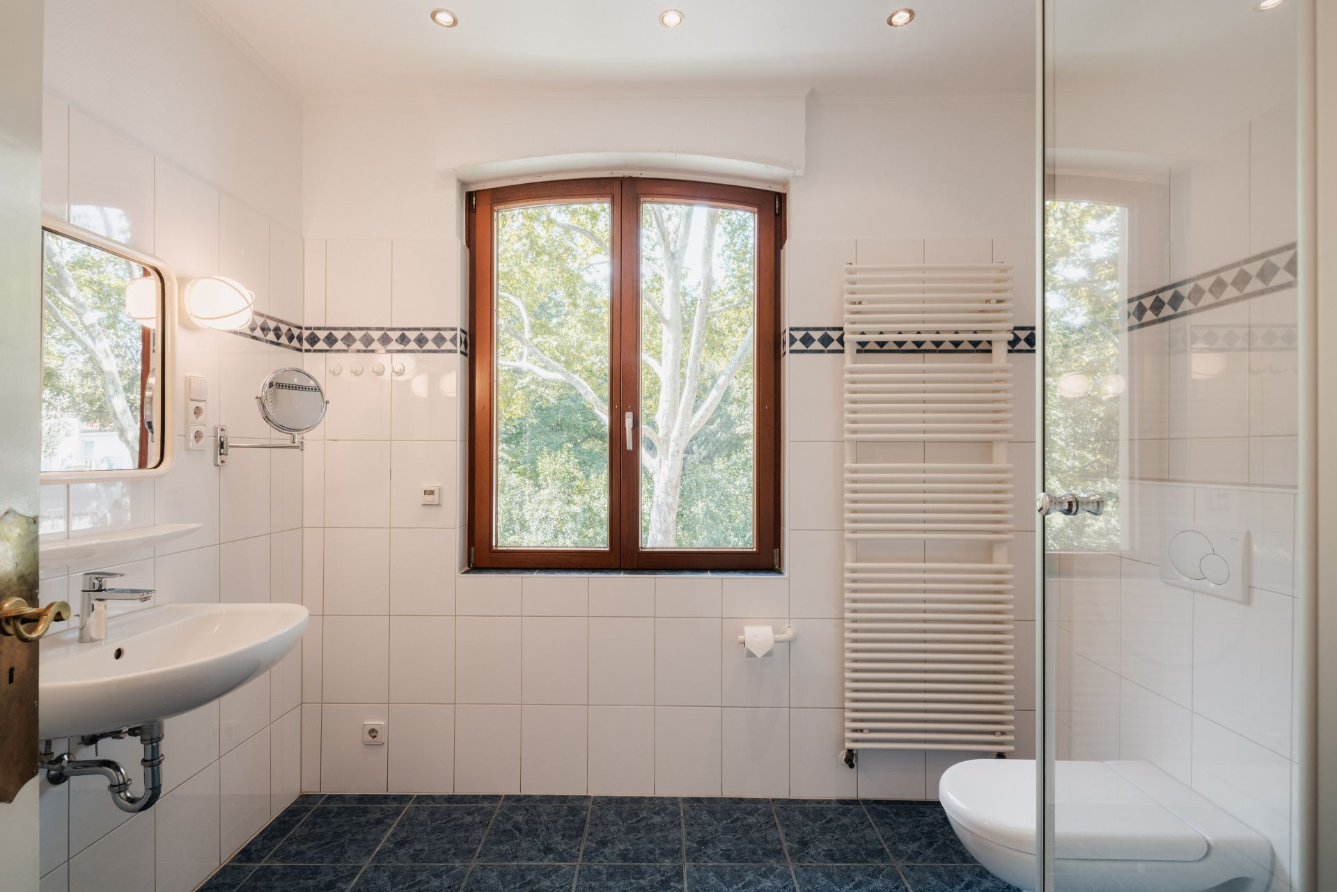 Helles Badezimmer mit Fenster, Waschbecken, Handtuchheizkörper und WC auf blauem Fliesenboden.