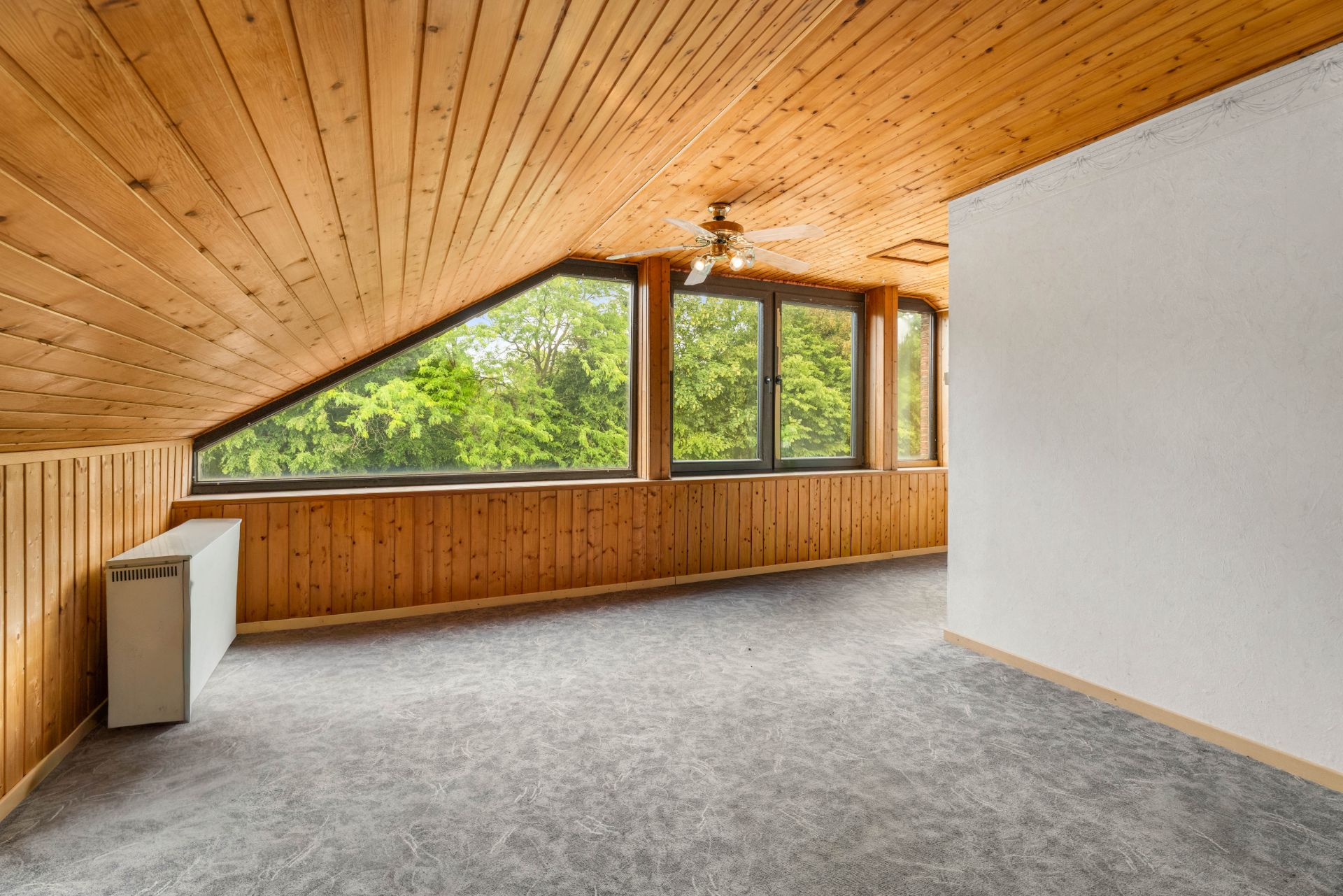 Dachzimmer mit Holzdecke, großen Fenstern und grauem Teppichboden, Blick ins Grüne.