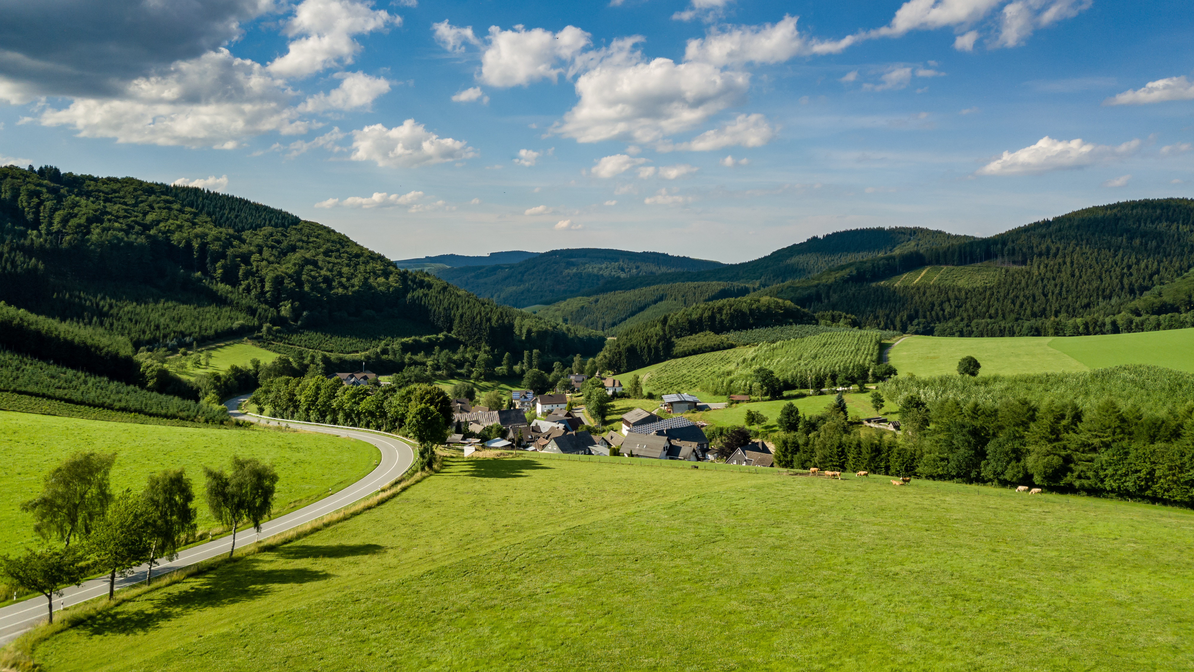 Dorf mit Häusern, umgeben von grünen Hügeln, Bäumen und einer Landstraße unter blauem Himmel.