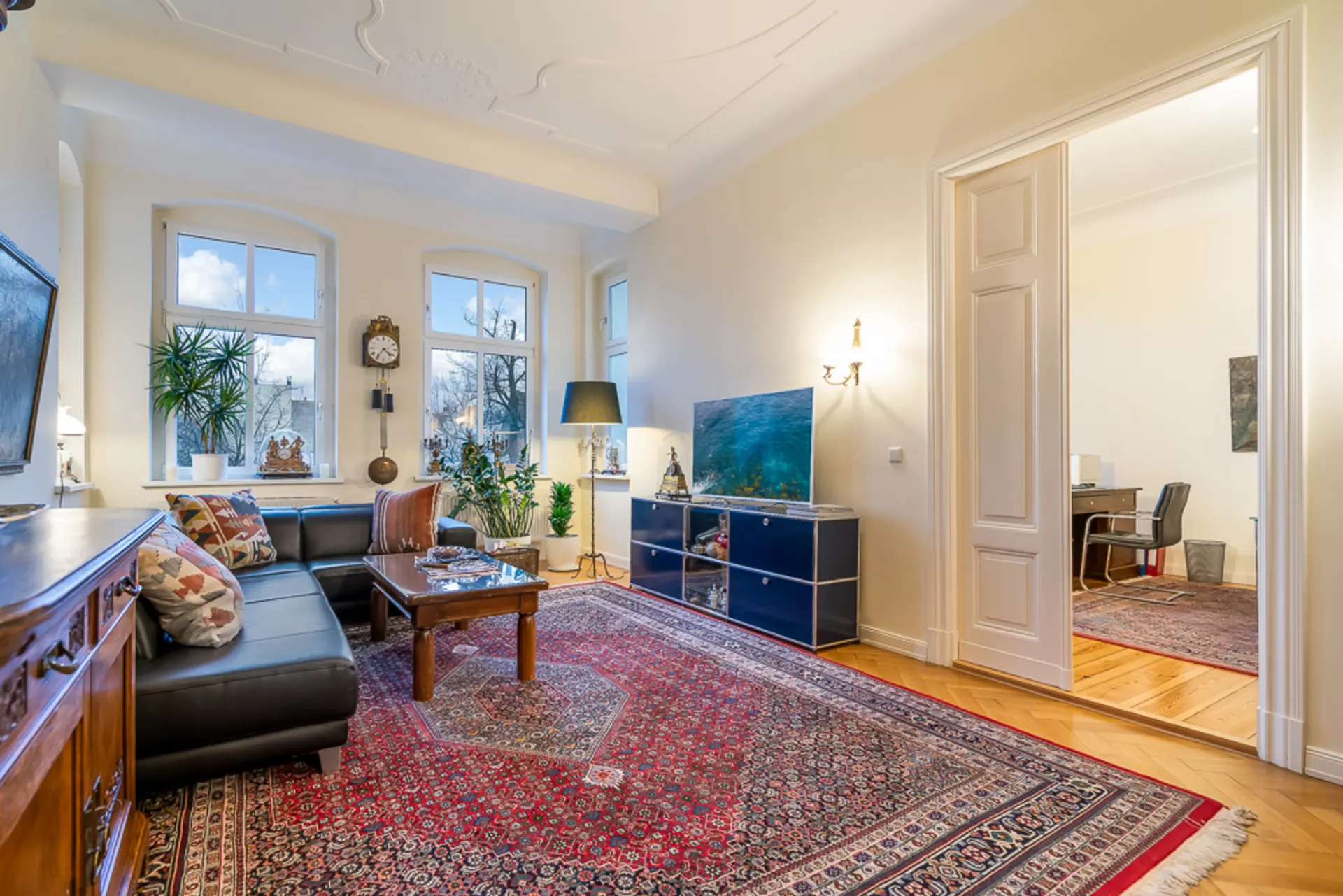 Helles Wohnzimmer mit Teppich, dunklem Sofa, Couchtisch, Pflanzen und großen Fenstern.