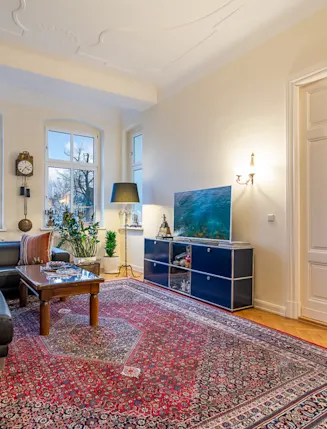 Helles Wohnzimmer mit Teppich, dunklem Sofa, Couchtisch, Pflanzen und großen Fenstern.