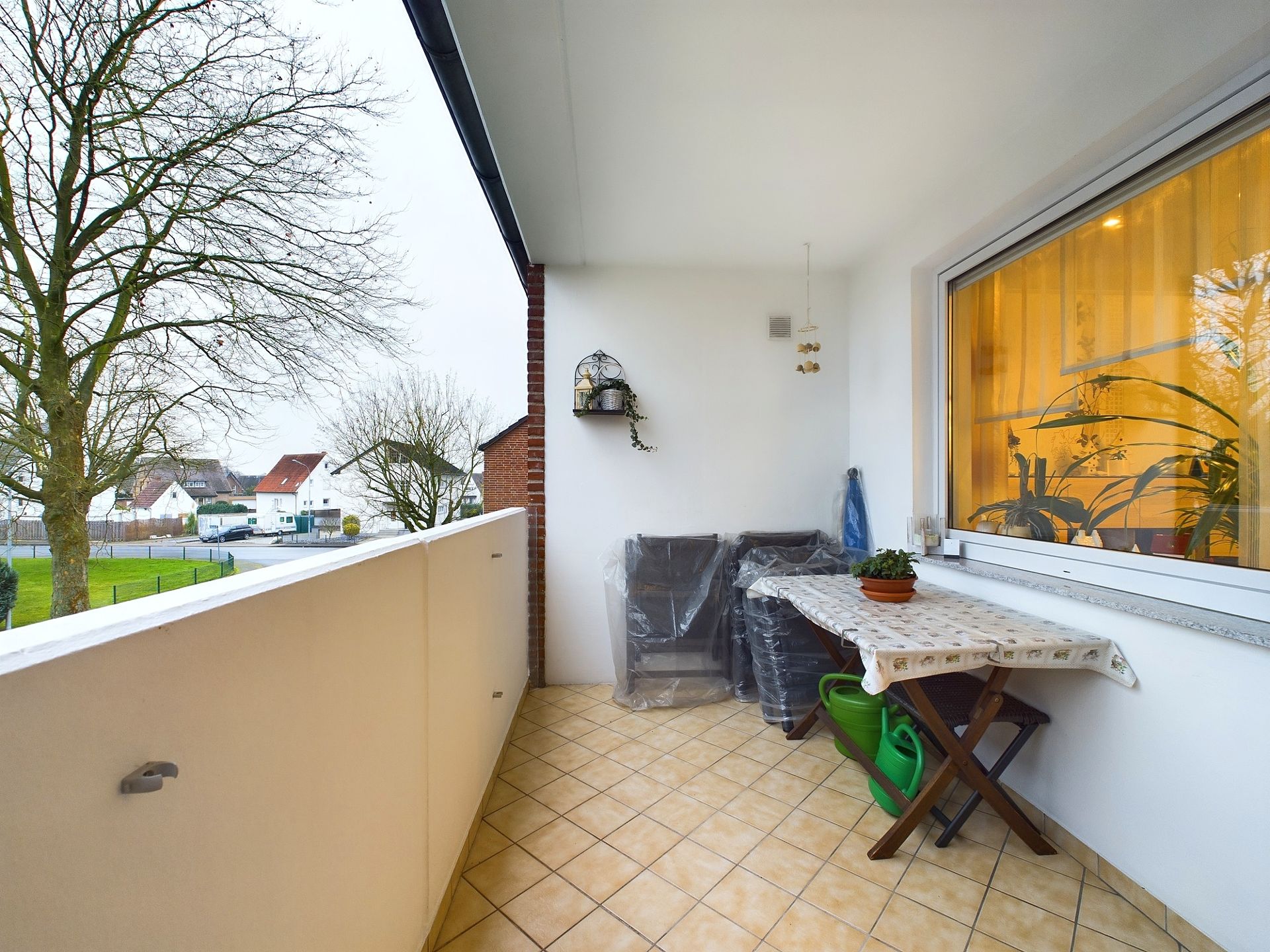 Überdachter Balkon mit Tisch, Stühlen, Pflanzen und Blick auf Bäume und Nachbarhäuser.