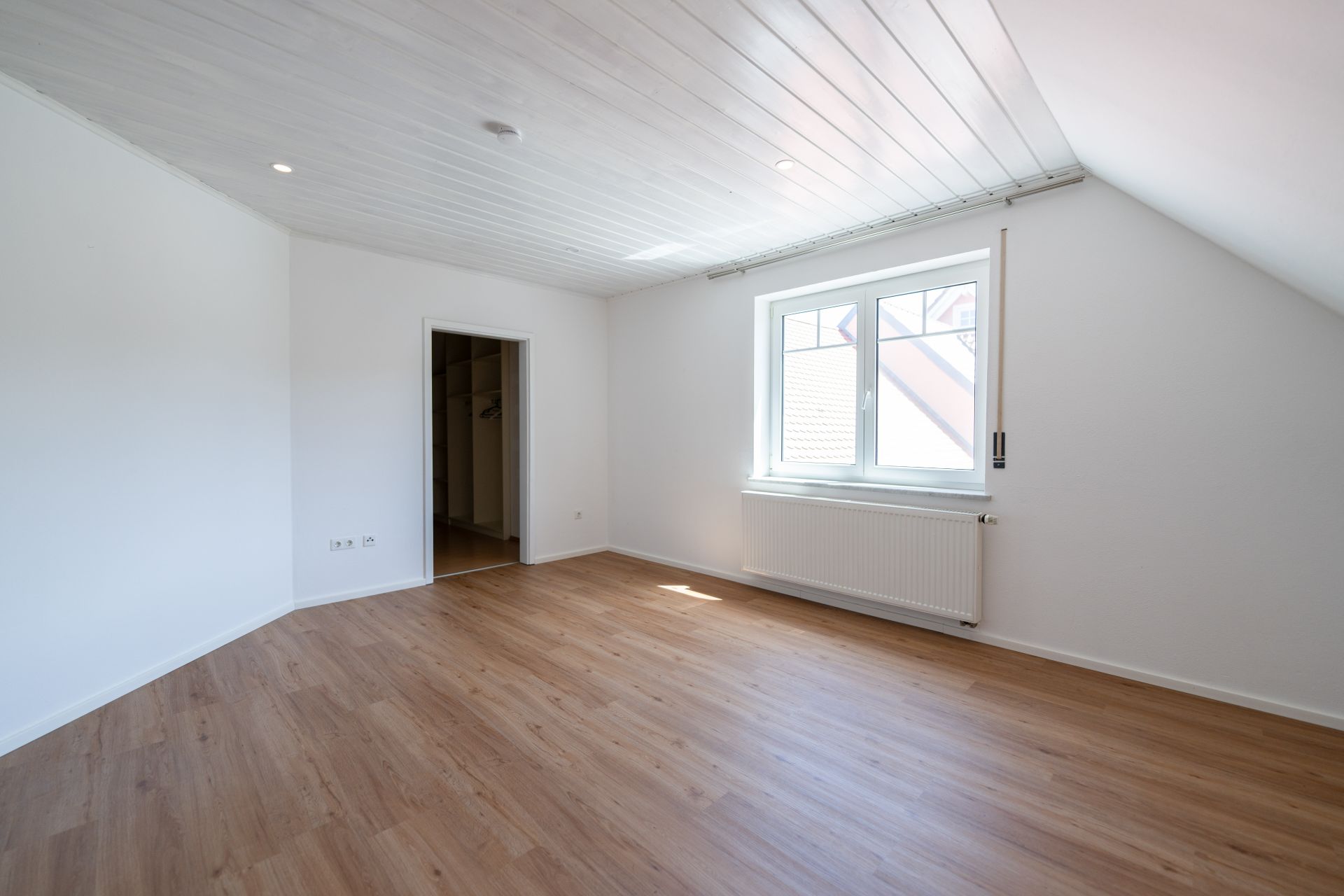 Helles, leeres Zimmer mit Holzfußboden, weißer Decke und großem Fenster.