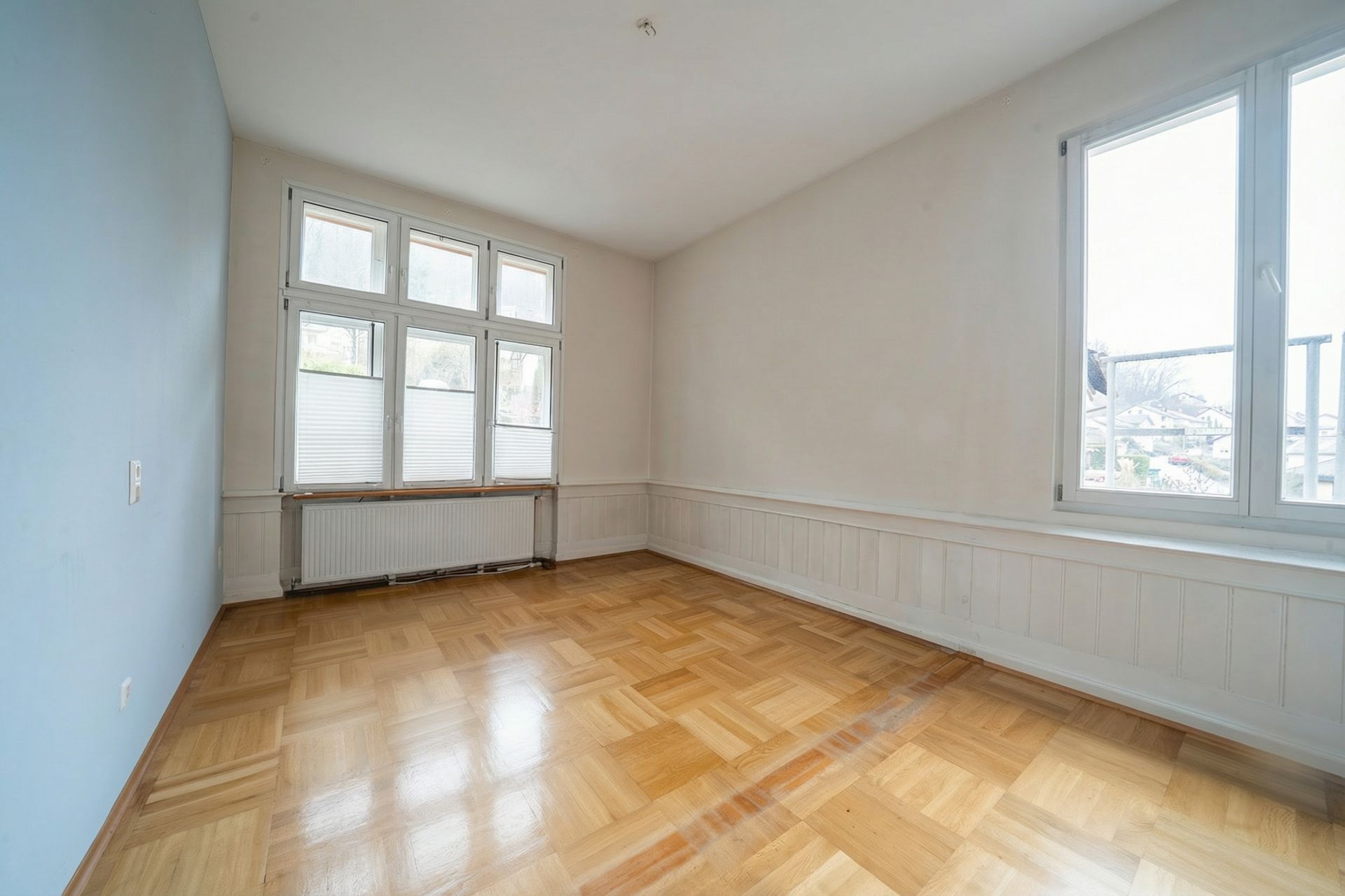 Leerer Raum mit Parkettboden, großen Fenstern und weiß gestrichenen Wänden.