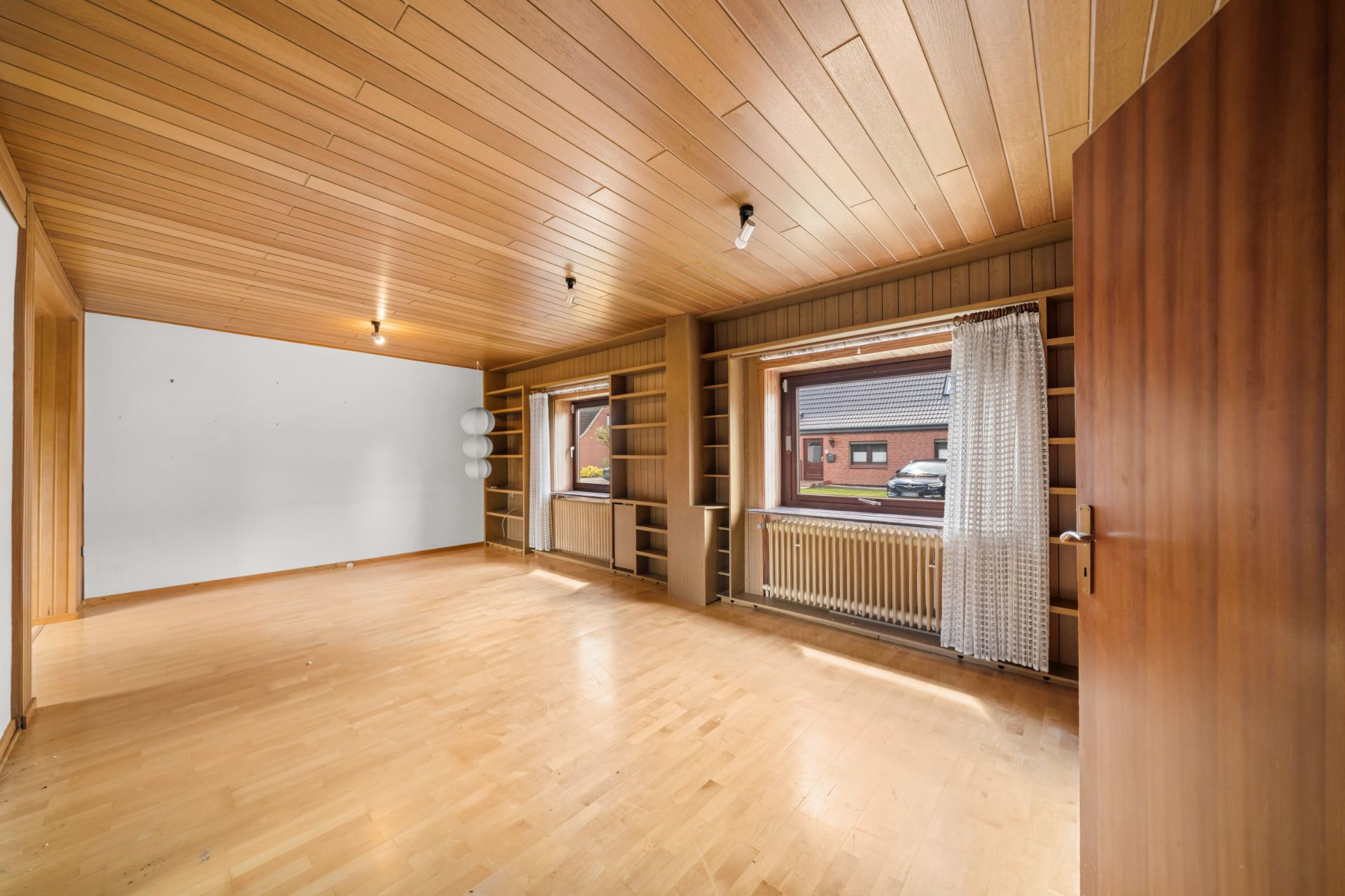 Helles Wohnzimmer mit Holzdecke, Holzfußboden, zwei großen Fenstern und offenen Regalen.