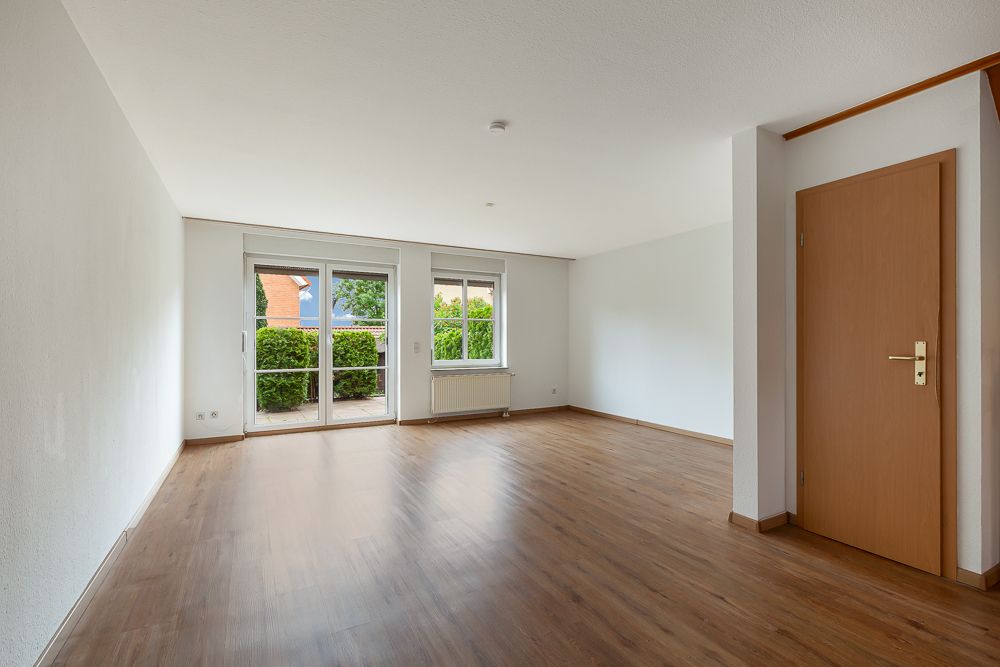 Helles, leeres Wohnzimmer mit Parkettboden, Fenster und Glastür zum Garten.