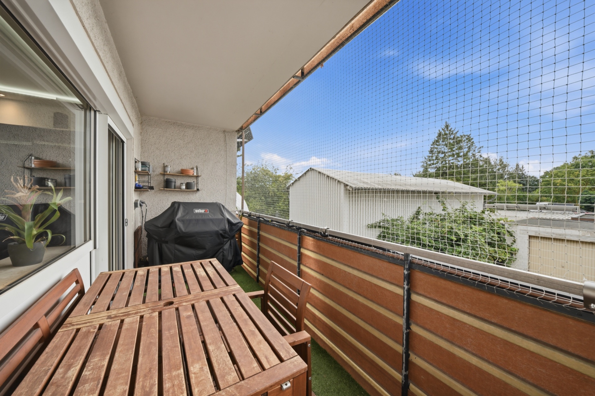 Balkon mit Holztisch, Stühlen, Grill und Pflanze, durch Netz gesichert, Ausblick auf Garten und Gebäude.