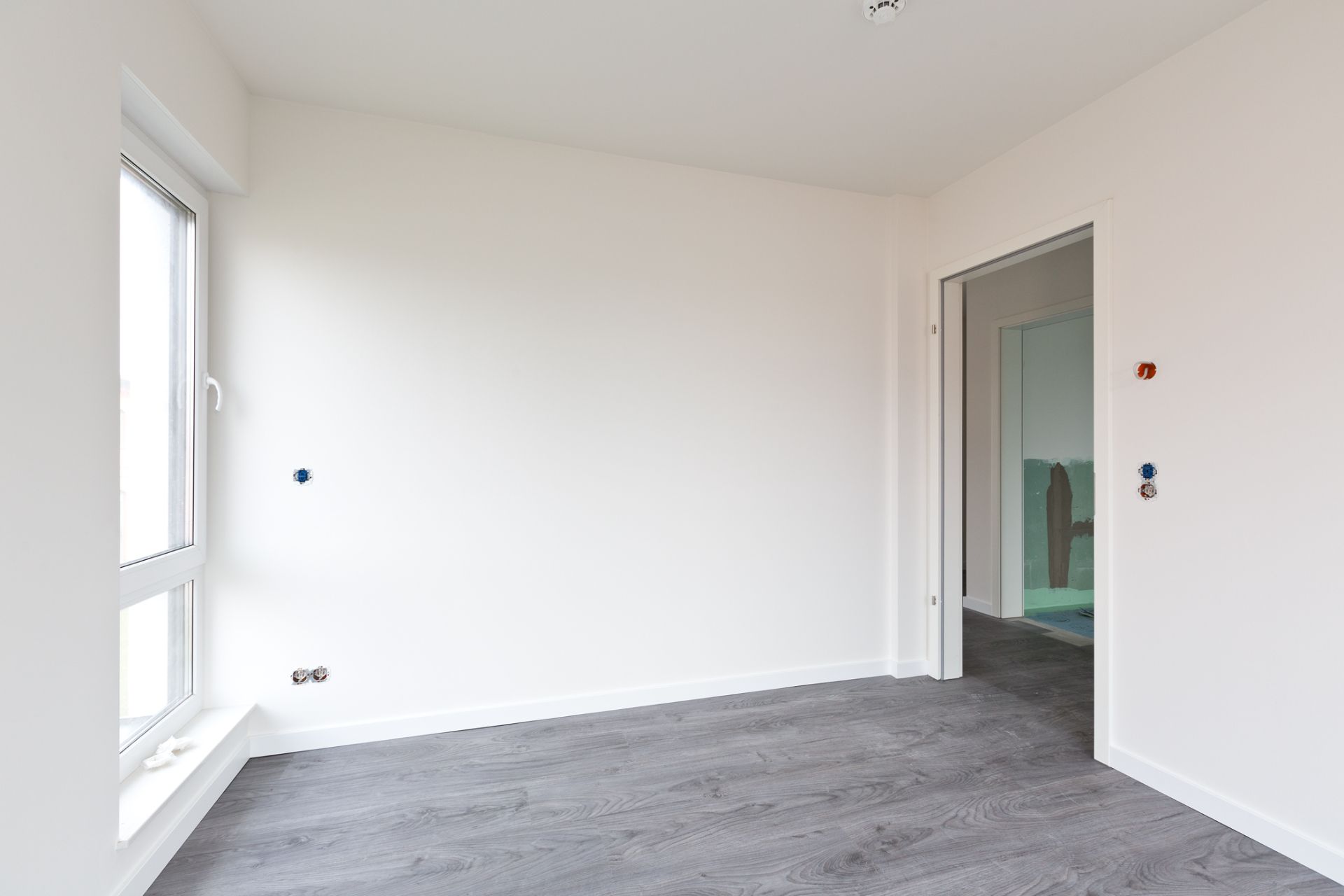 Leerer Raum mit grauem Boden und weißer Wand, Fenster links, offene Tür rechts.