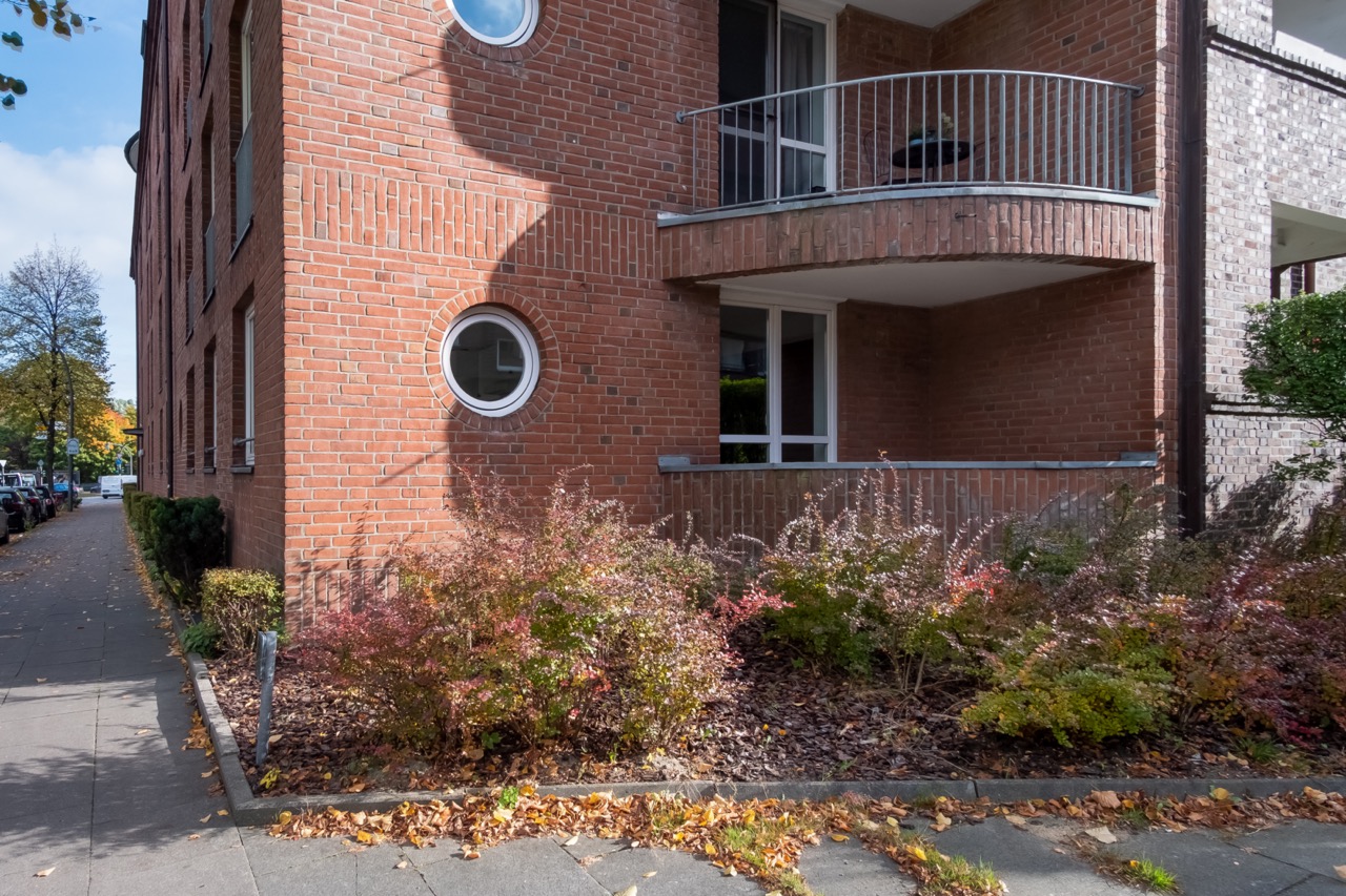 Erdgeschosswohnung mit rotem Backstein, runder Fensterform und überdachtem Balkon, umgeben von Sträuchern.