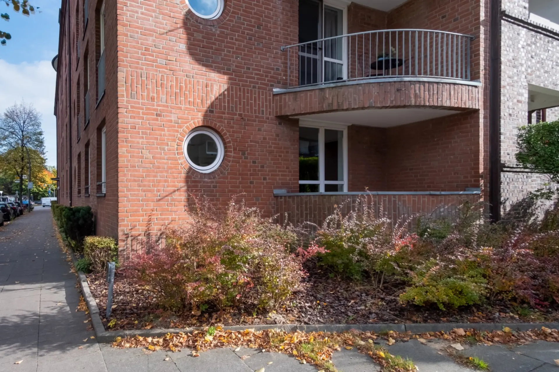 Erdgeschosswohnung mit rotem Backstein, runder Fensterform und überdachtem Balkon, umgeben von Sträuchern.