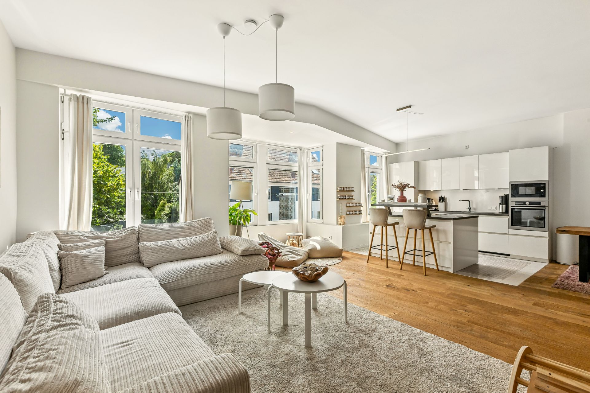 Helles Wohnzimmer mit Ecksofa, Teppich, offener Küche und großen Fenstern.