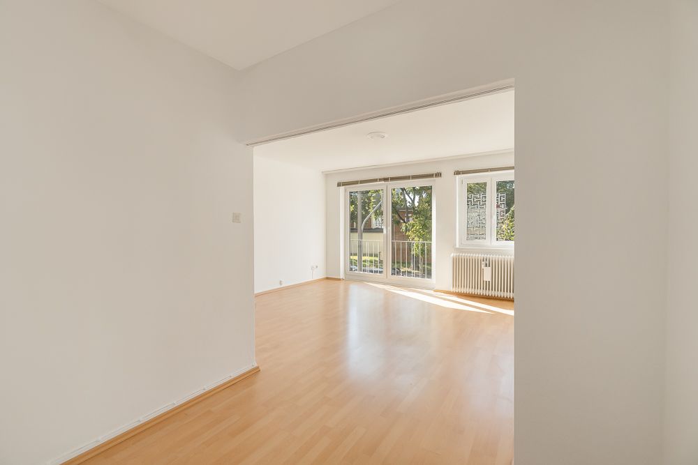 Helles, leeres Wohnzimmer mit Parkettboden und großen Fenstern mit Blick nach draußen.