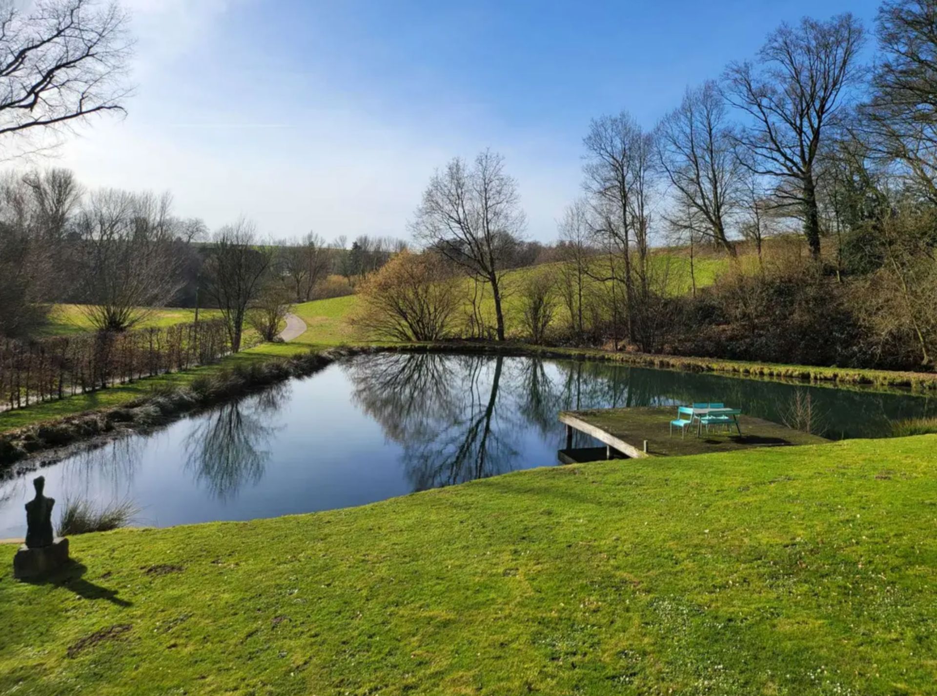 Teich mit Steg und Sitzgruppe auf einer grünen Wiese, im Hintergrund Bäume und blauer Himmel.