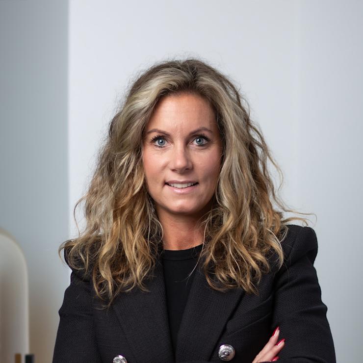 Frau mit lockigen, blonden Haaren in schwarzem Blazer mit verschränkten Armen vor weißer Wand.