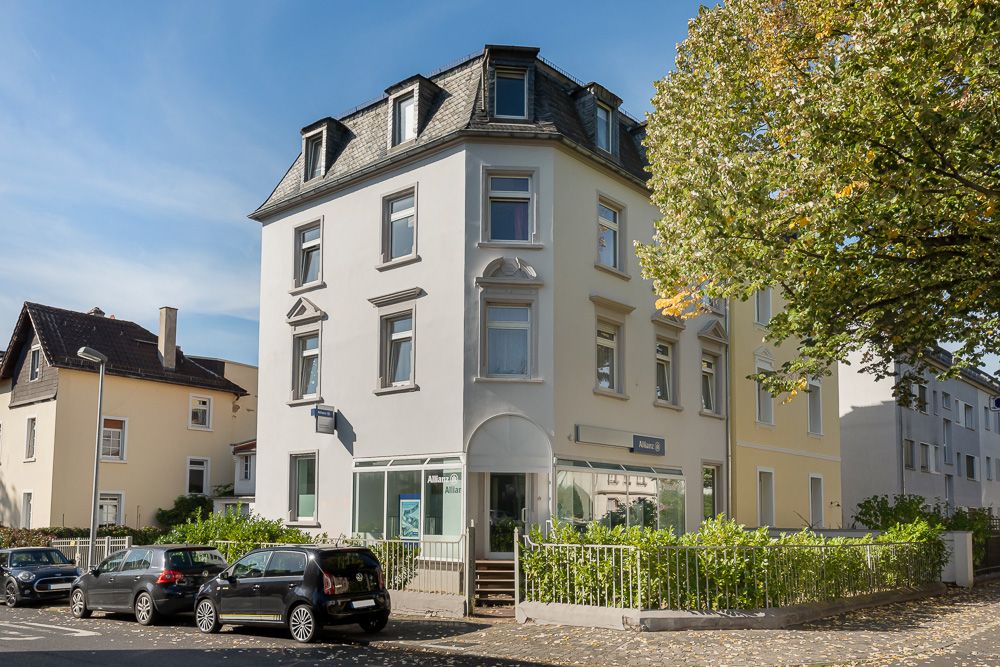Weißes, dreistöckiges Wohnhaus mit Mansarddach und mehreren Autos davor an einer Straßenecke.