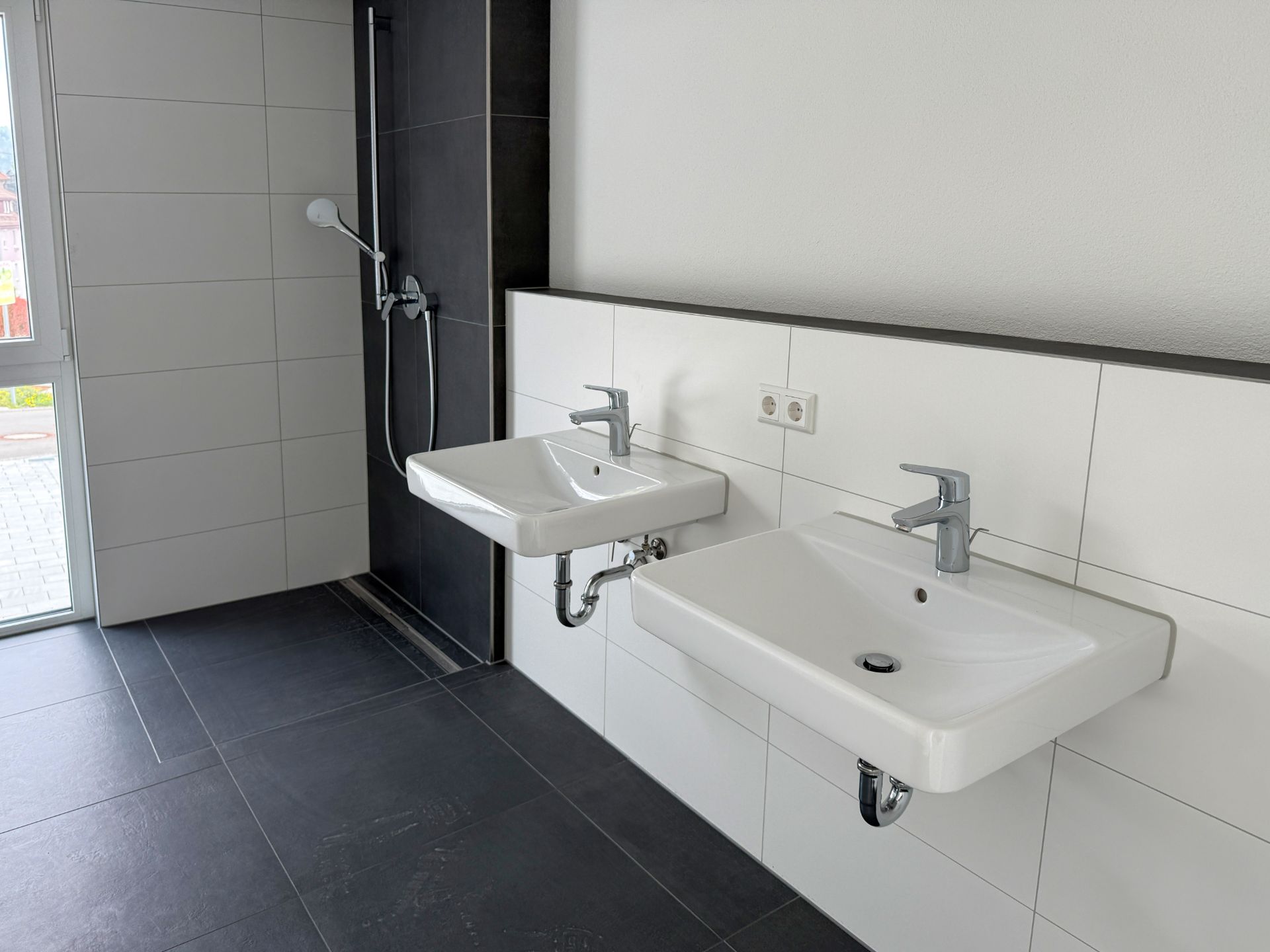 Modernes Bad mit zwei Waschbecken und bodengleicher Dusche, weiße Wandfliesen, dunkle Bodenfliesen.