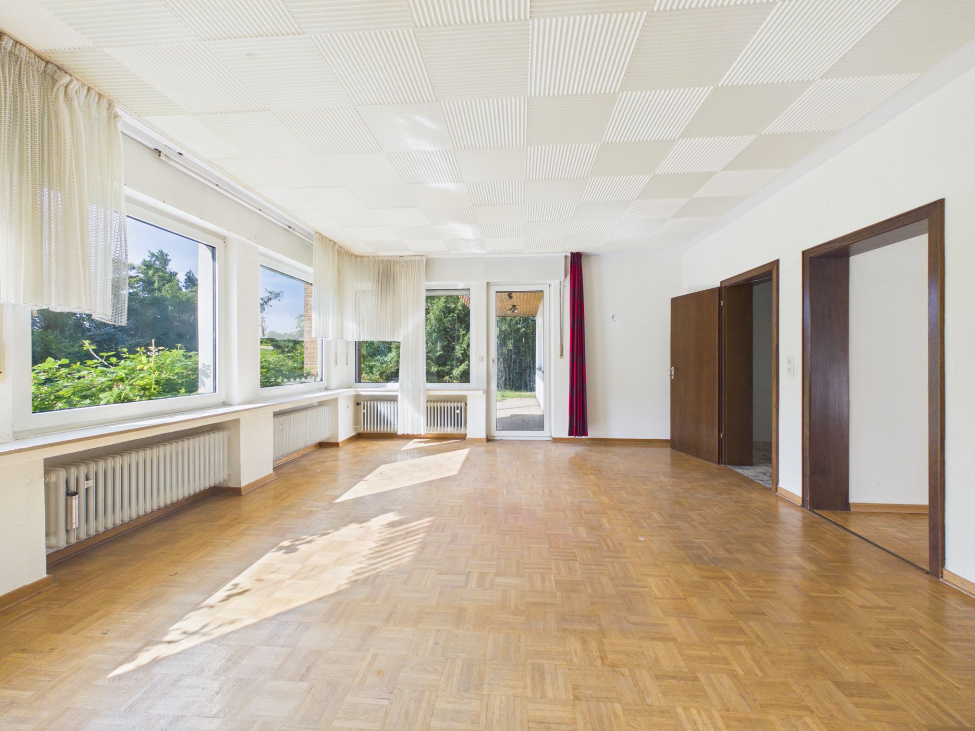 Heller, leerer Raum mit Parkettboden, großen Fenstern und weißen Vorhängen.