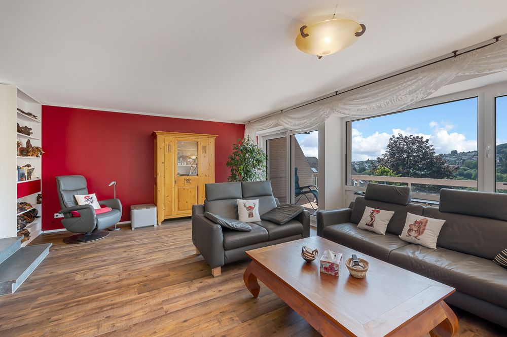 Wohnzimmer mit zwei grauen Sofas, Sessel, Holztisch, Schrank und großen Fenstern mit Ausblick.