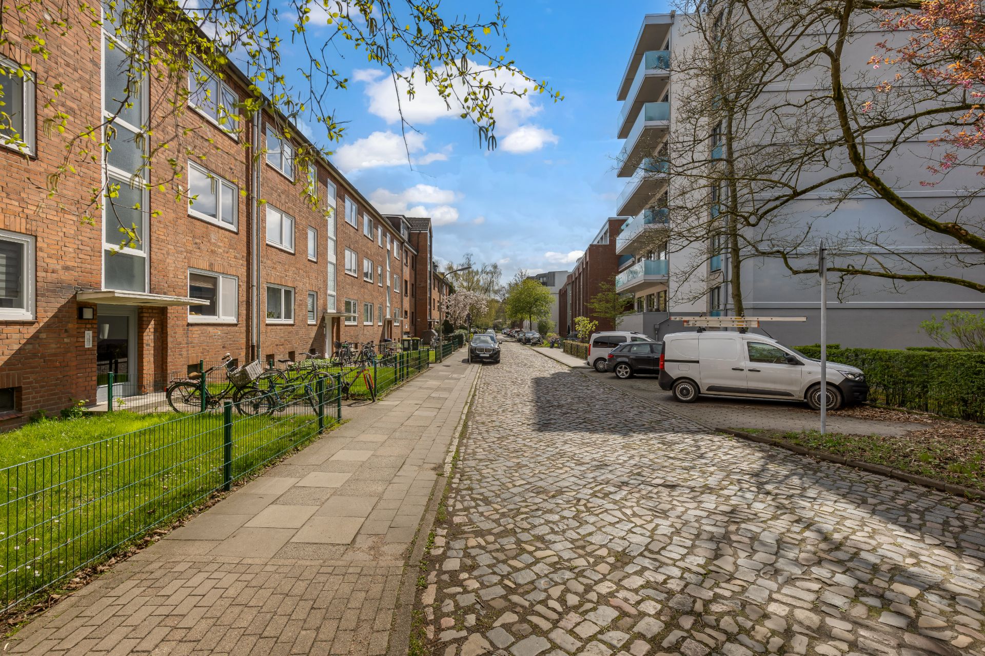 Gepflasterte Wohnstraße mit parkenden Autos, Fahrrädern und Mehrfamilienhäusern bei sonnigem Wetter.