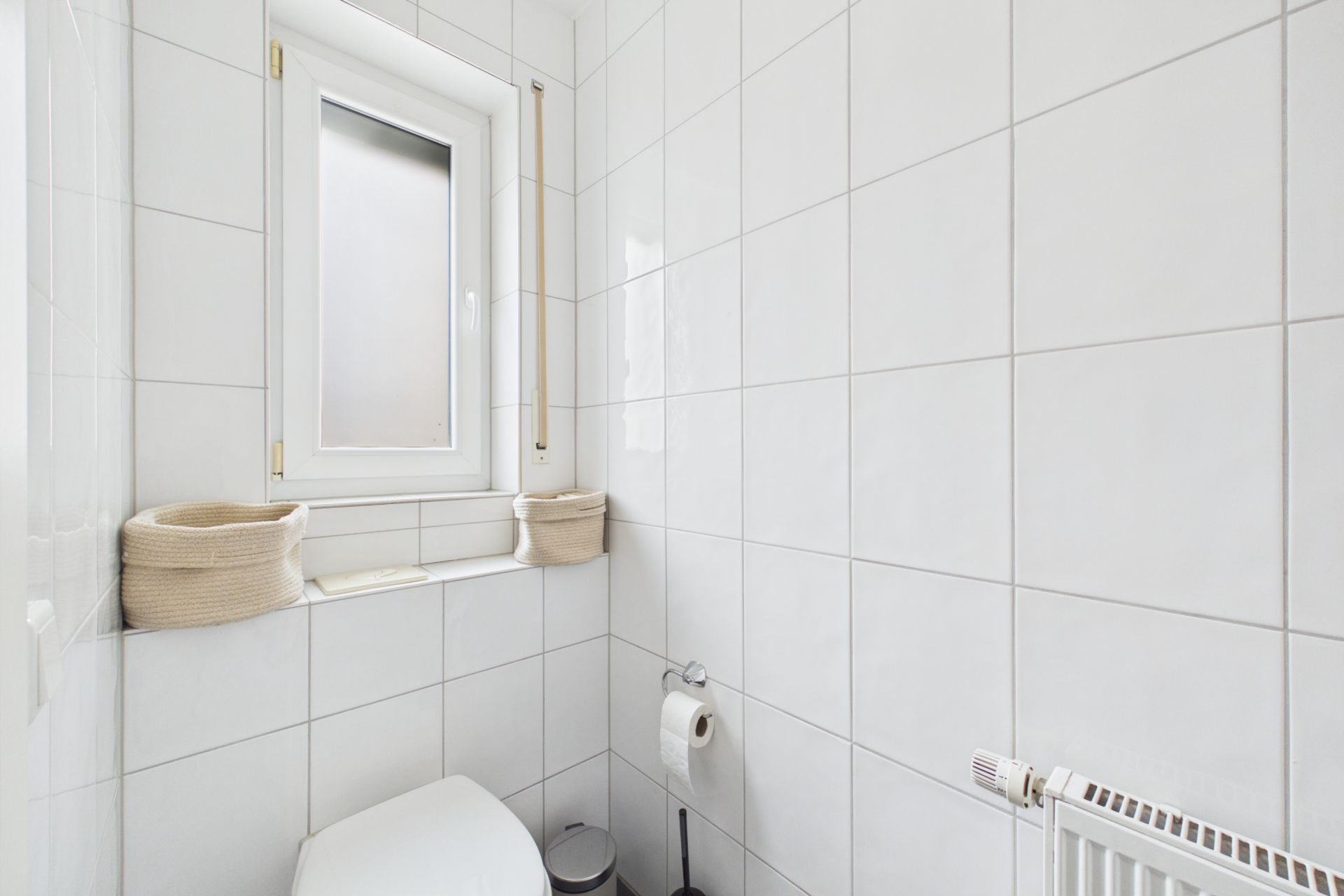 Kleines, weiß gefliestes Bad mit Fenster, Toilette, Heizung und zwei Körben auf der Fensterbank.
