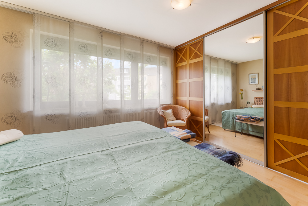 Schlafzimmer mit grünem Bett, großem Fenster, Spiegelkleiderschrank und Sessel.