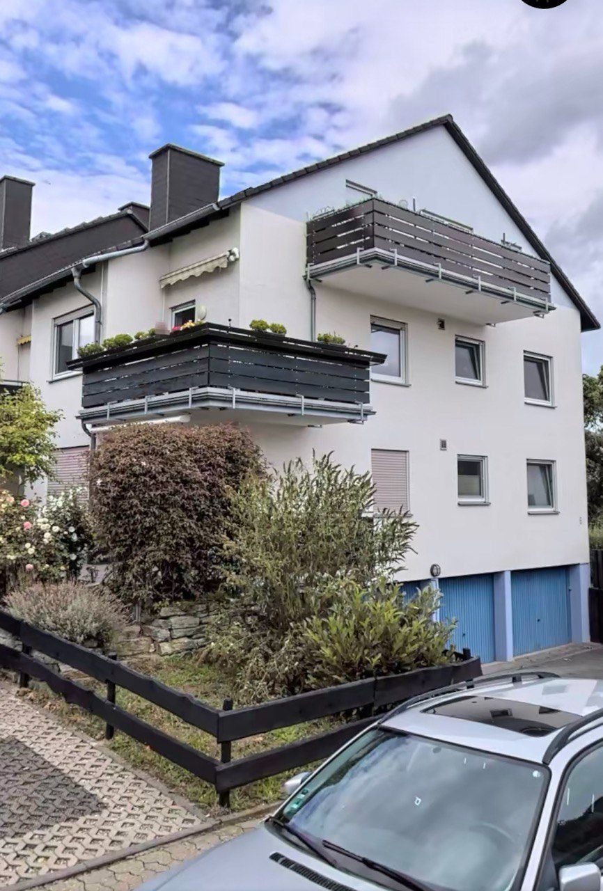 Wohnhaus mit zwei Balkonen, Garten, blauen Garagentoren und geparktem Auto im Vordergrund.